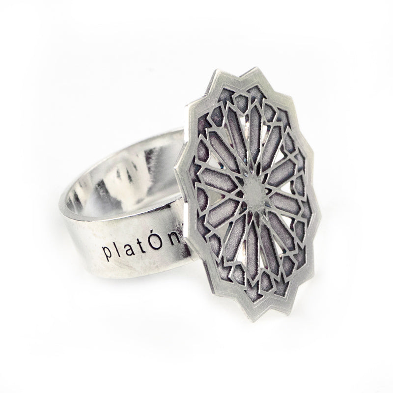 The Silver Ring Estrella Big by Platónica is a sterling silver adjustable piece featuring a geometric starburst or rose window motif on a large flat disc. Handmade in Granada, the band is engraved with platÓn, offering an exquisite contemporary jewelry design.