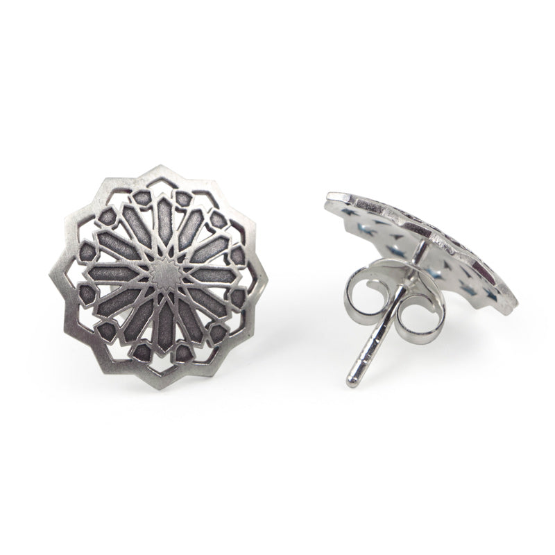 Part of Platónicas Andalusí collection, the Sterling Silver Studs Earrings Estrella feature intricate geometric designs like a spoked wheel or star. One earring is shown upright to emphasize its front detail, while the other lies flat to display its handcrafted stud and back closure.