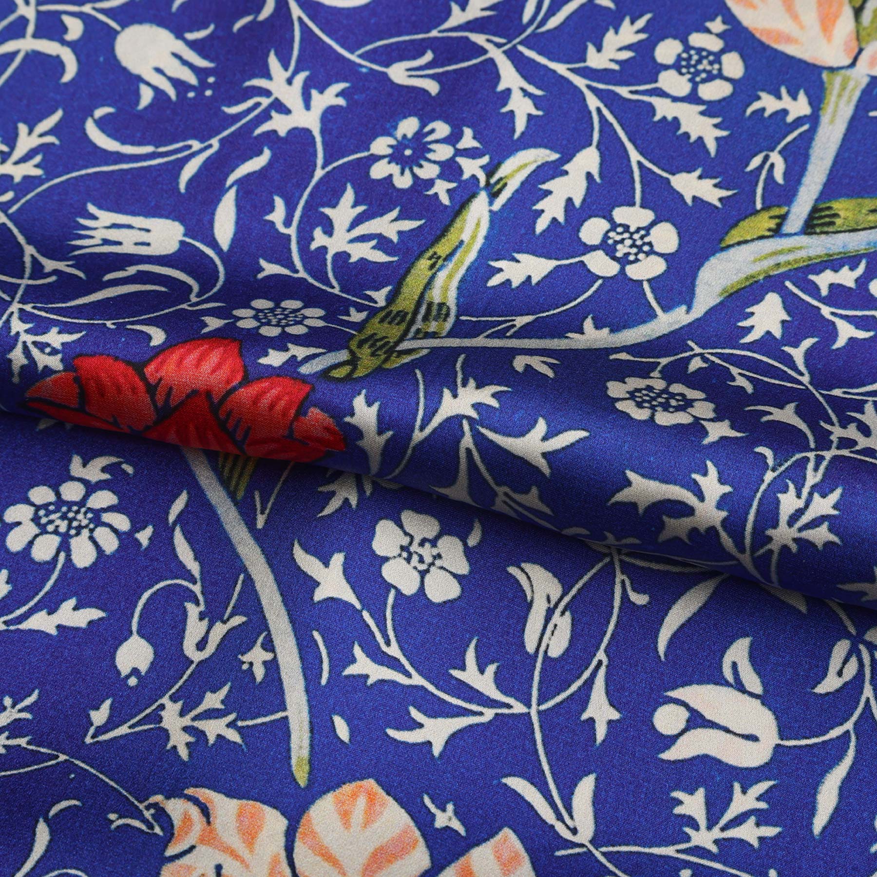 A close-up of the Floral Blue Silk Scarf Galata: luxurious blue silk with white floral patterns, bold red and pink Art Nouveau flowers, and a red-edged hem, gently draped on a white surface.
