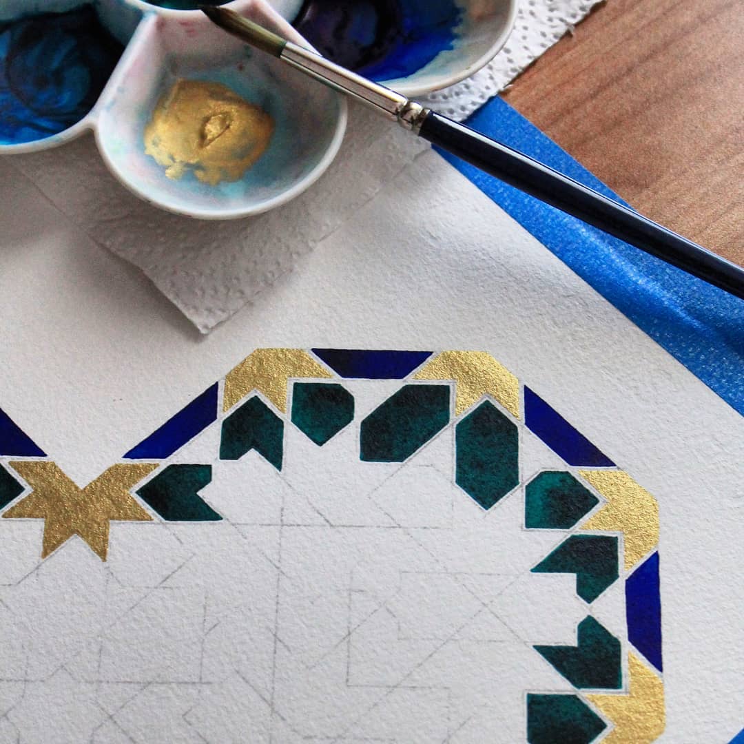 A close-up of a geometric art piece in progress, featuring gold, teal, and blue shapes. A paintbrush rests on a palette with blue and gold paint, and part of the design is outlined on textured white paper.