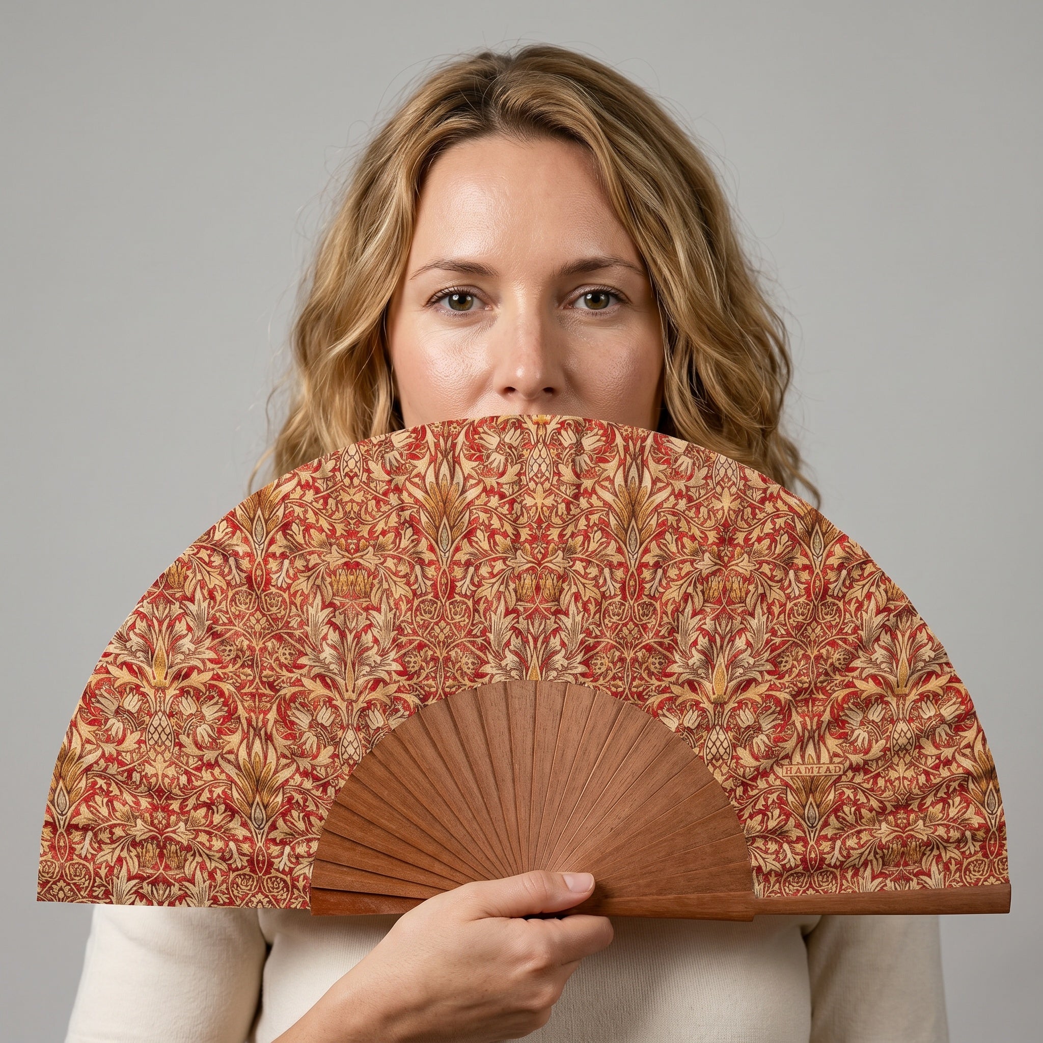 A woman holding the William Morris Print Hand Fan Renacentista, with its William Morris botanical print in red and gold, against a soft neutral background.