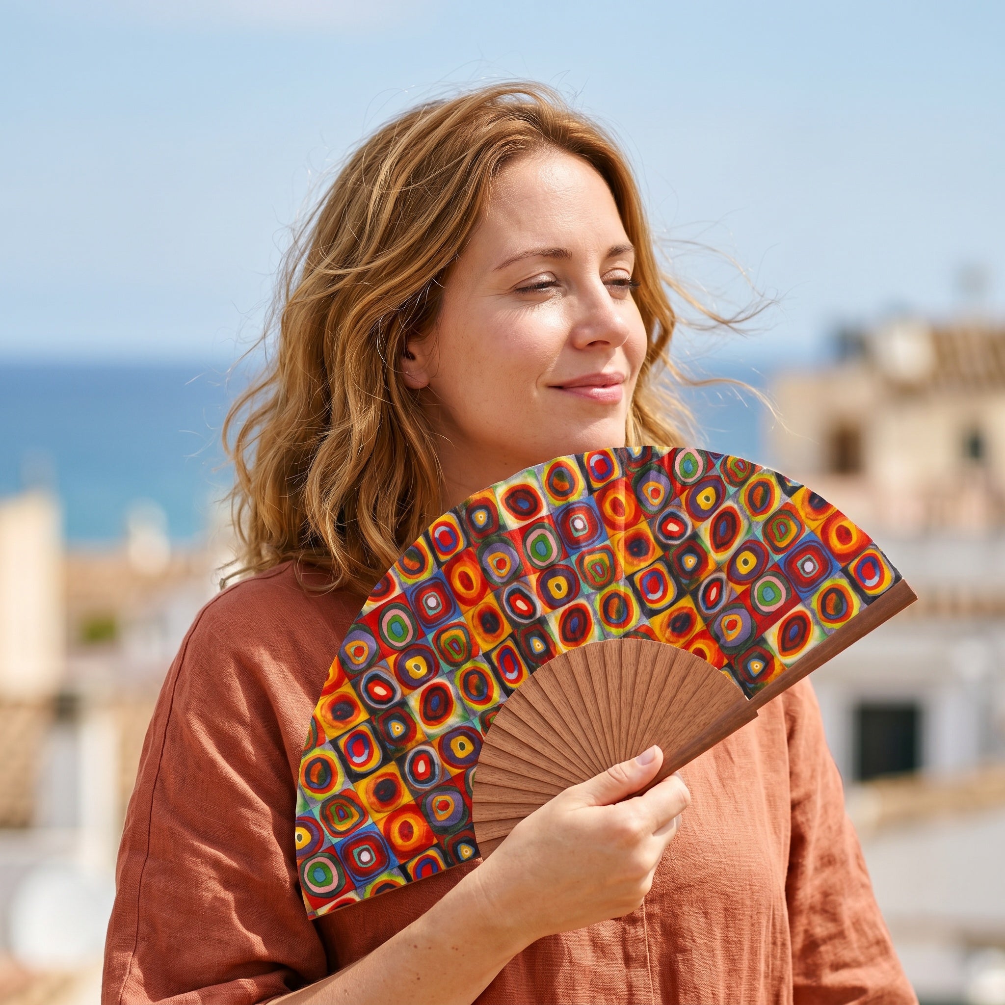 A woman holding the Kandinsky Print Hand Fan Lorelei, with its Kandinsky concentric circles colourful print, against a soft neutral background.