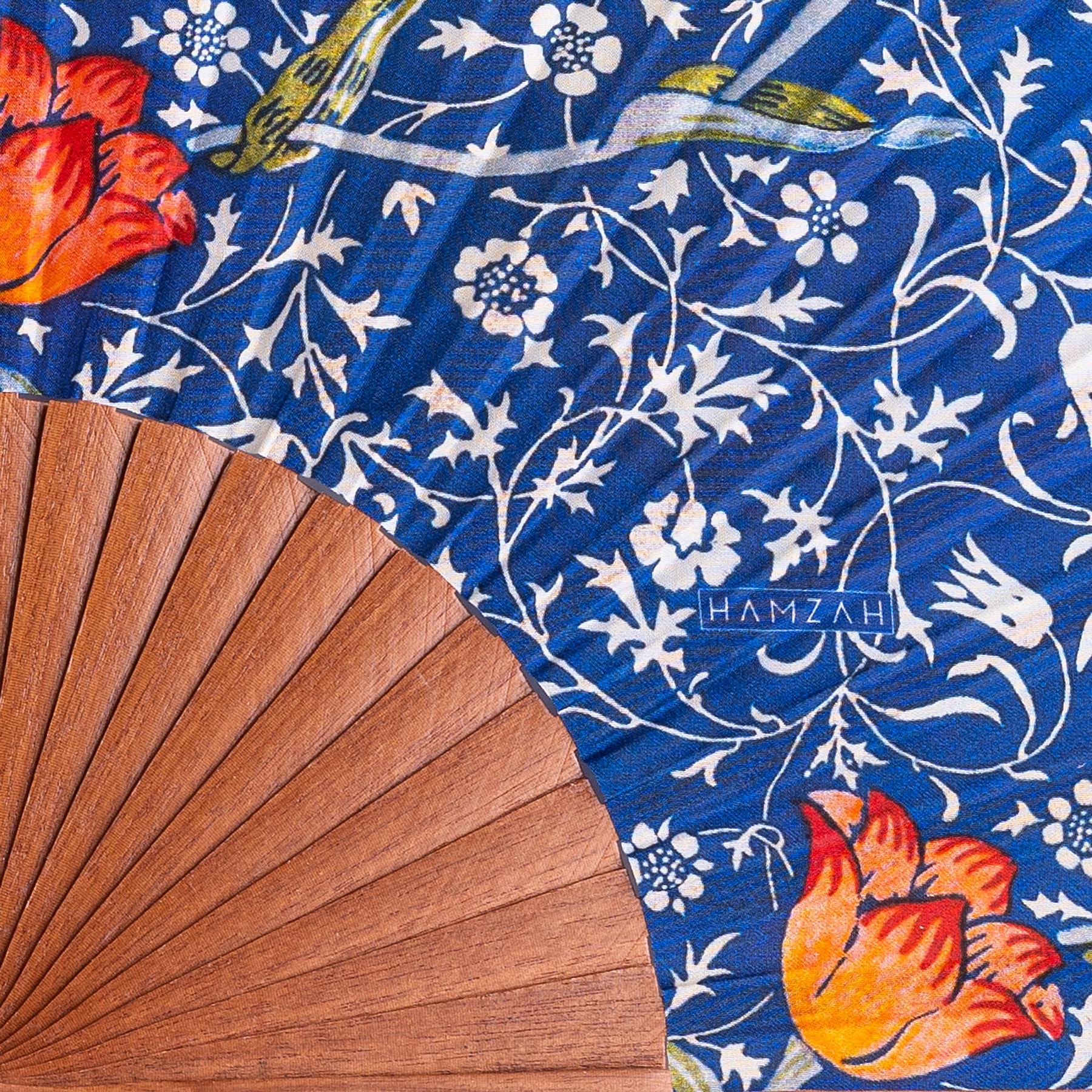 The Natural Silk Fan W. Morris Print Gálata rests on a blue floral fabric with red-orange flowers, green stems, and white blossoms. "HAMZAH" appears in white letters on the fabric.
