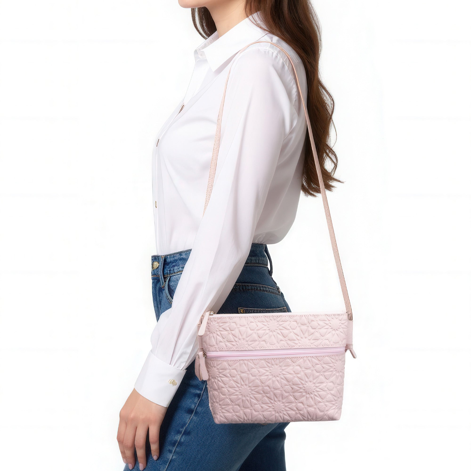A woman in a white button-up shirt and blue jeans carries a Pale Pink Embossed Crossbody Bag with an Islamic Design Pattern, featuring a zipper closure and long strap, elegantly worn over her shoulder.