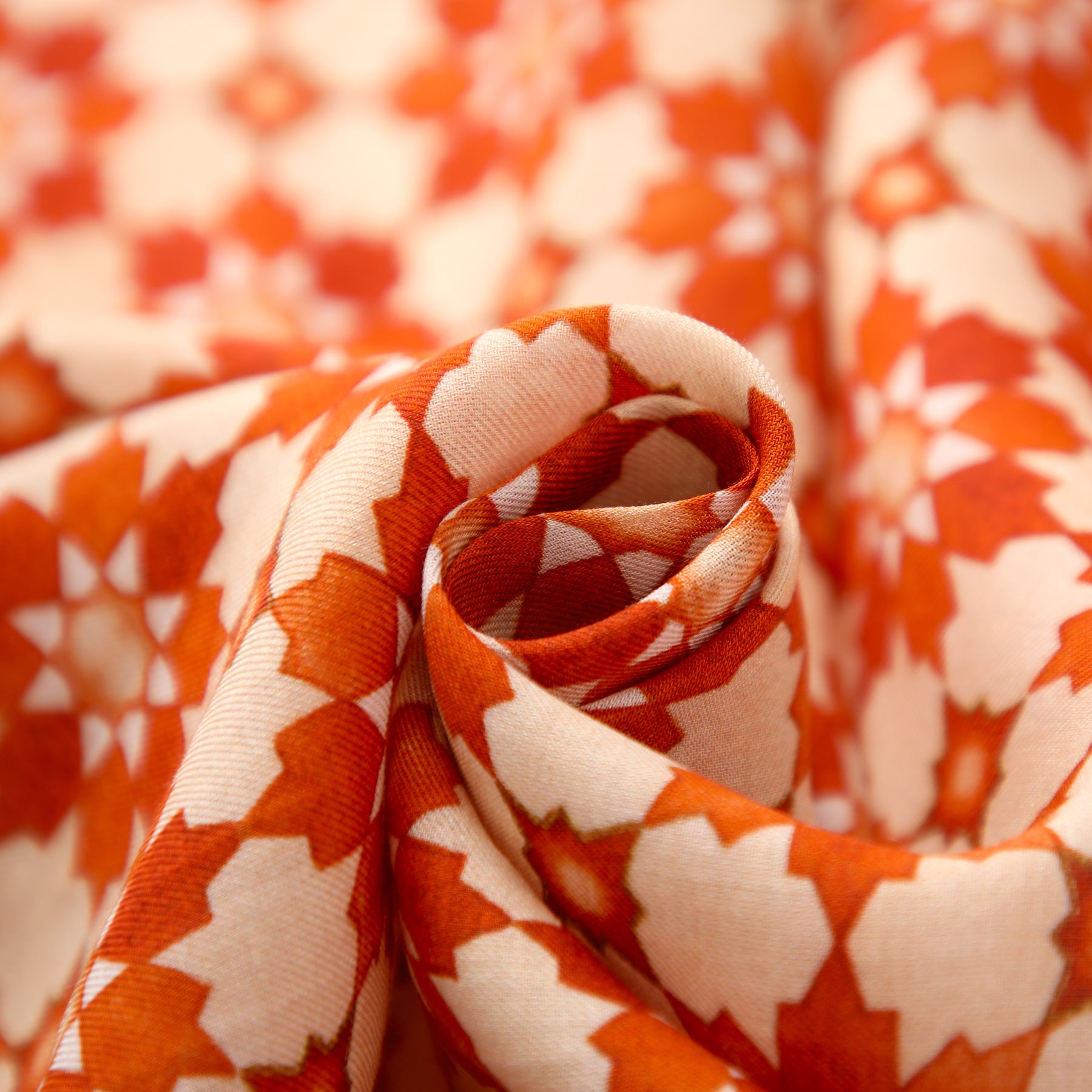 A close-up of the Orange Scarf with Islamic Pattern Print, softly folded in a swirl, reveals its Alhambra-inspired geometric star motif on white patterned fabric.