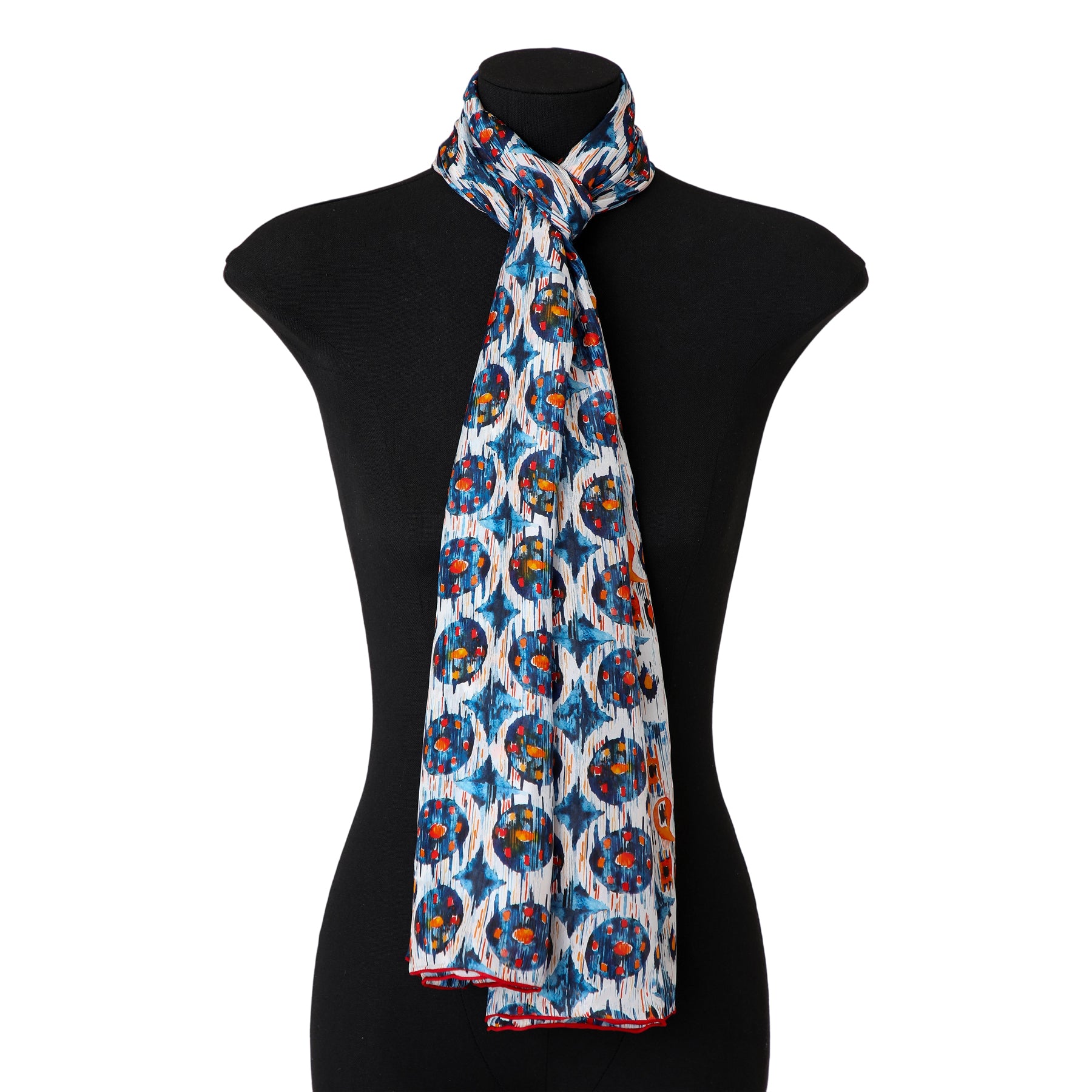The White and Blue Habotai Silk Scarf Ikat Print, featuring blue, white, red, and yellow circular ethnic designs, is draped around a black mannequin bust against a white background.