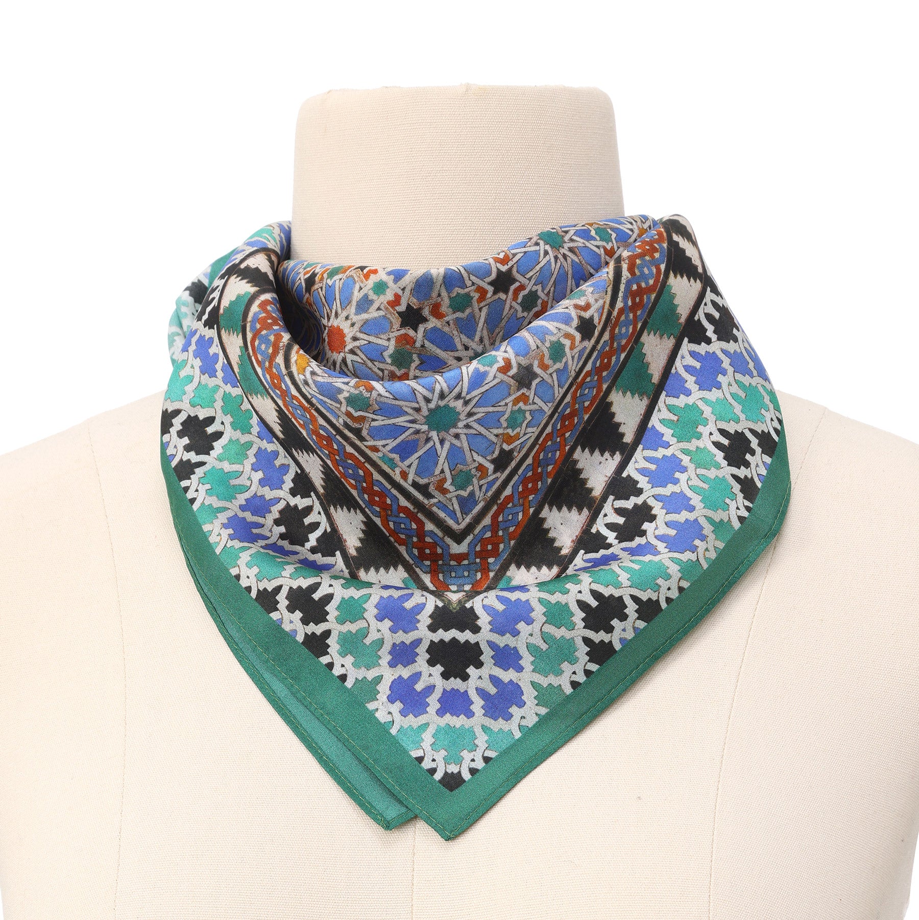 A mannequin displays the Alhambra Art Silk Square Scarf, featuring intricate geometric and mosaic patterns inspired by Islamic art in blue, green, black, orange, and white, folded into a triangle and wrapped around the neck.