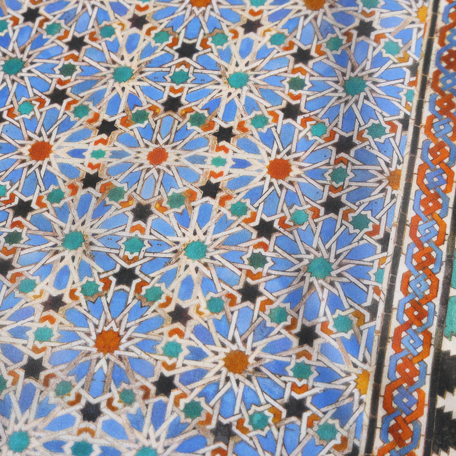 The Alhambra Art Silk Square Scarf features blue and white geometric star motifs with orange accents and a detailed border, capturing the elegance of Islamic art and the intricate craftsmanship of Spanish design.