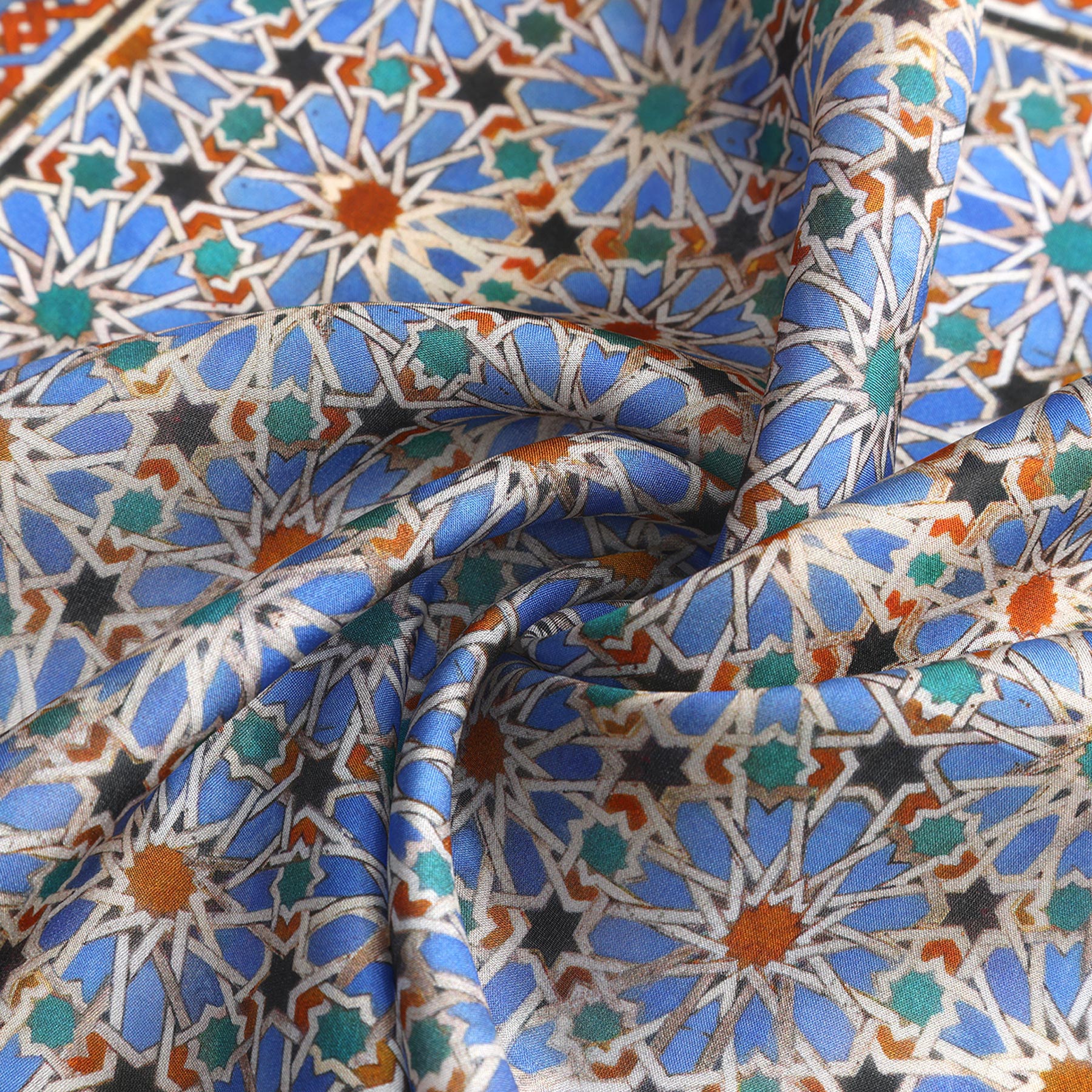 A close-up of the Alhambra Art Silk Square Scarf shows its intricate geometric patterns in blue, white, green, brown, and orange—star shapes and interlocking motifs inspired by Spanish craftsmanship—softly gathered in elegant folds.