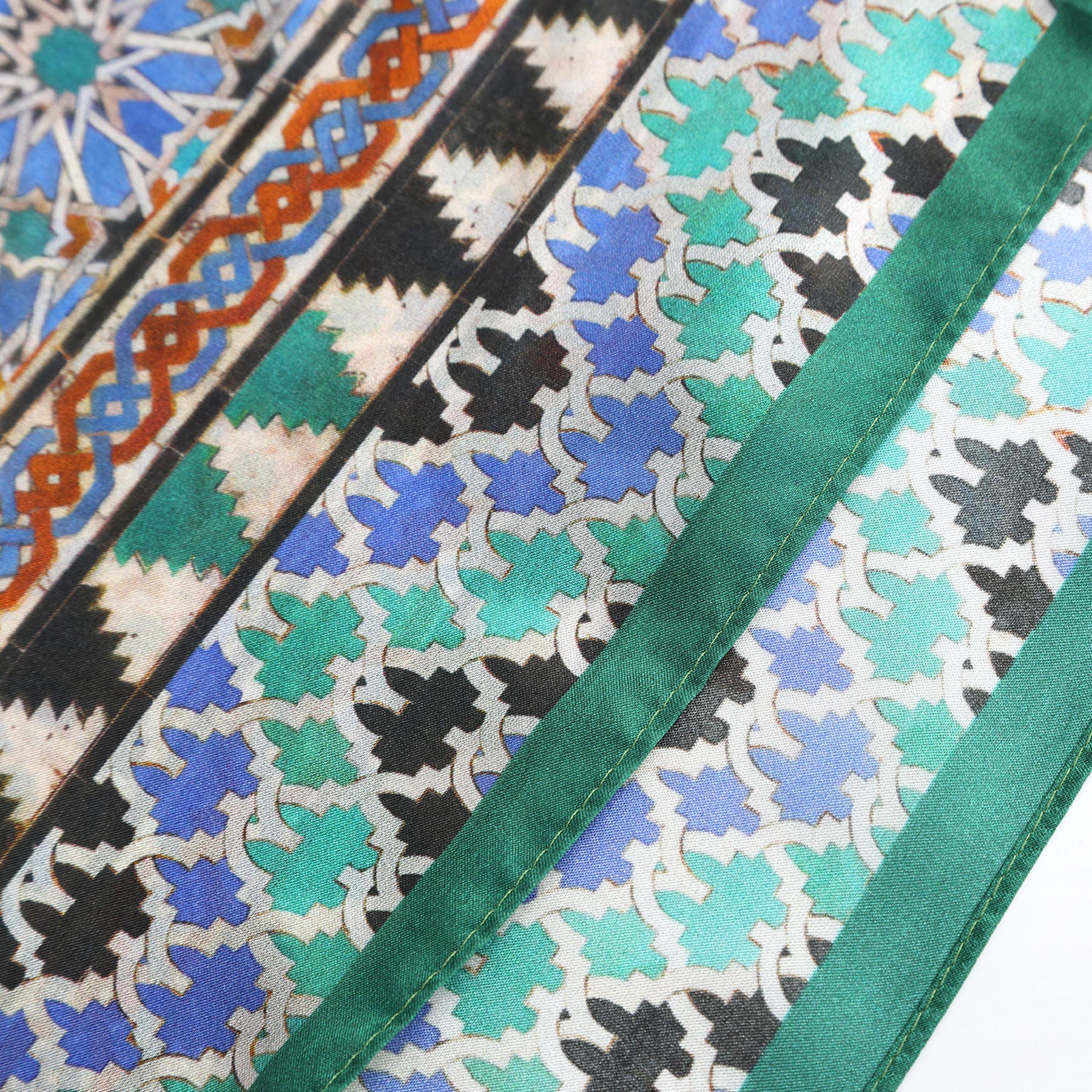 A close-up of the Alhambra Art Silk Square Scarf shows intricate blue, green, white, and black geometric patterns with a green border, inspired by traditional Islamic art and Spanish craftsmanship.