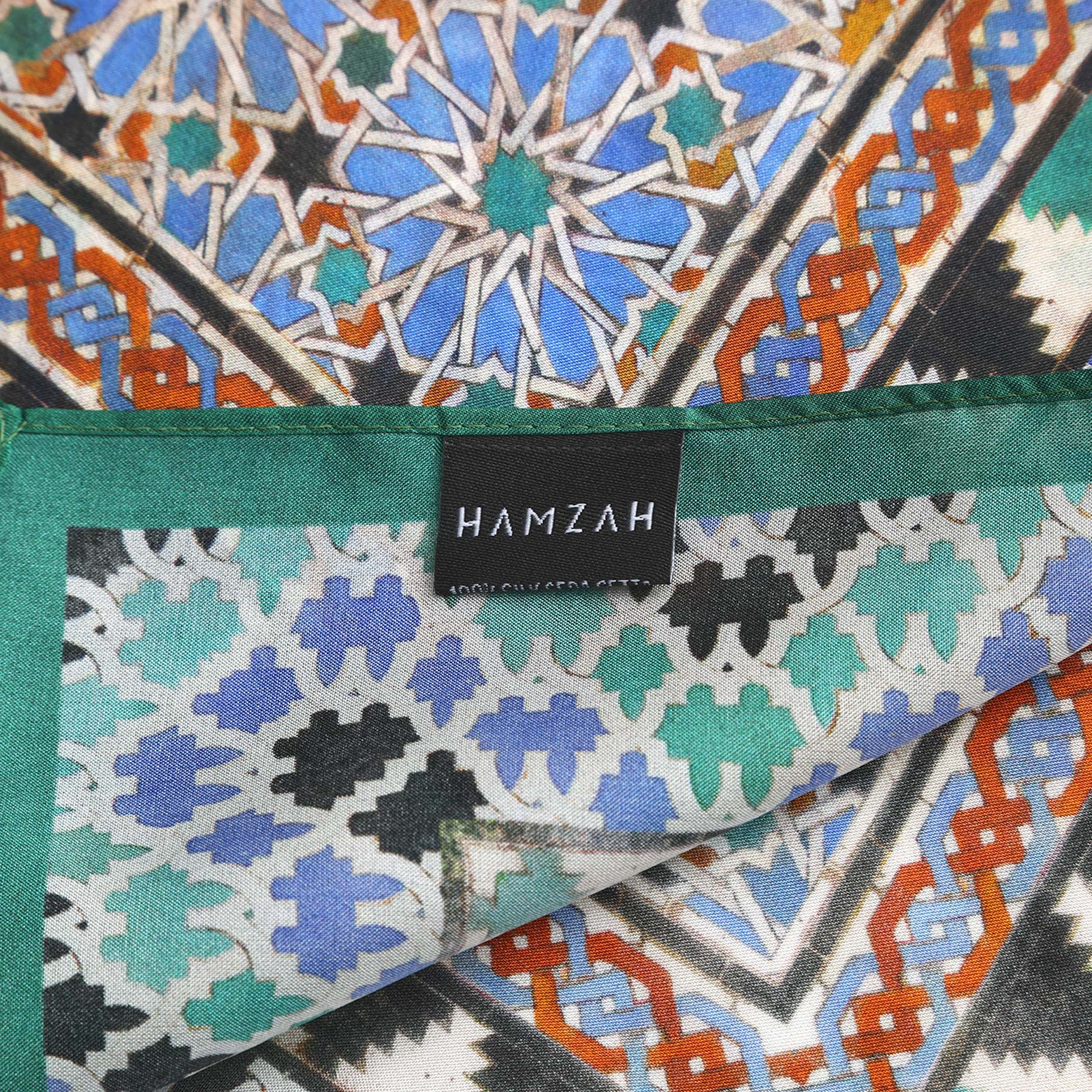A close-up of the Alhambra Art Silk Square Scarf showcases its vibrant blue, green, orange, and white geometric patterns and a black HAMZAH tag sewn on the edge.