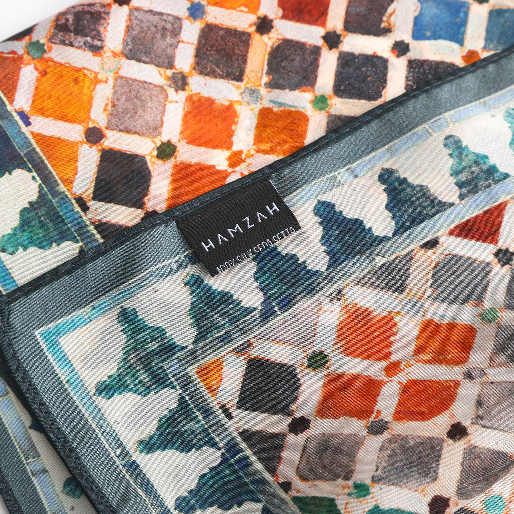 A close-up of the Colorful Square Scarf Comares, featuring a vibrant geometric Alhambra-inspired pattern in orange, blue, and white. A black label reads HAMZAH 100% SILK / MADE IN ITALY.