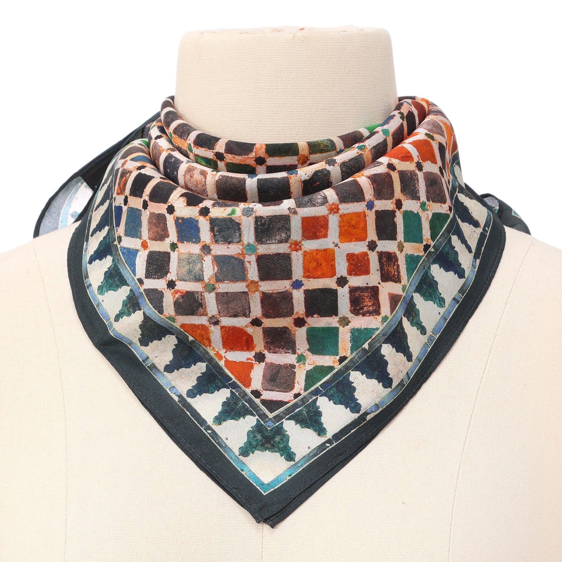 A mannequin showcases the Colorful Square Scarf Comares, a silk square scarf with a geometric checkered pattern inspired by Alhambra Palace Islamic art in orange, brown, green, and blue hues. It features black and teal borders and is loosely tied around the neck.