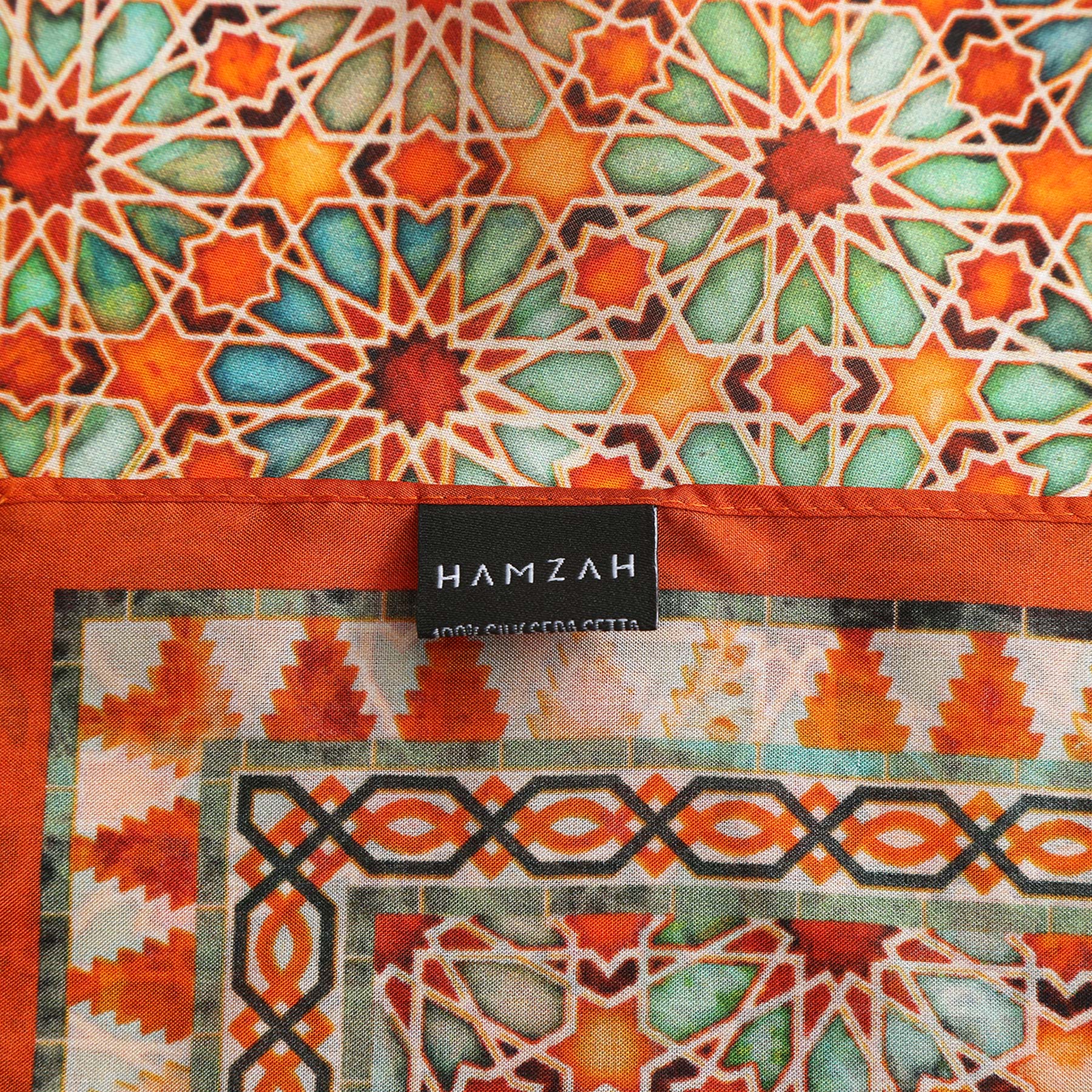 The Orange Square Scarf Zellige features intricate Islamic-inspired geometric patterns in orange, green, and cream. A black label reading "HAMZAH 100% SUTERA CETAK" is sewn onto the fabric.