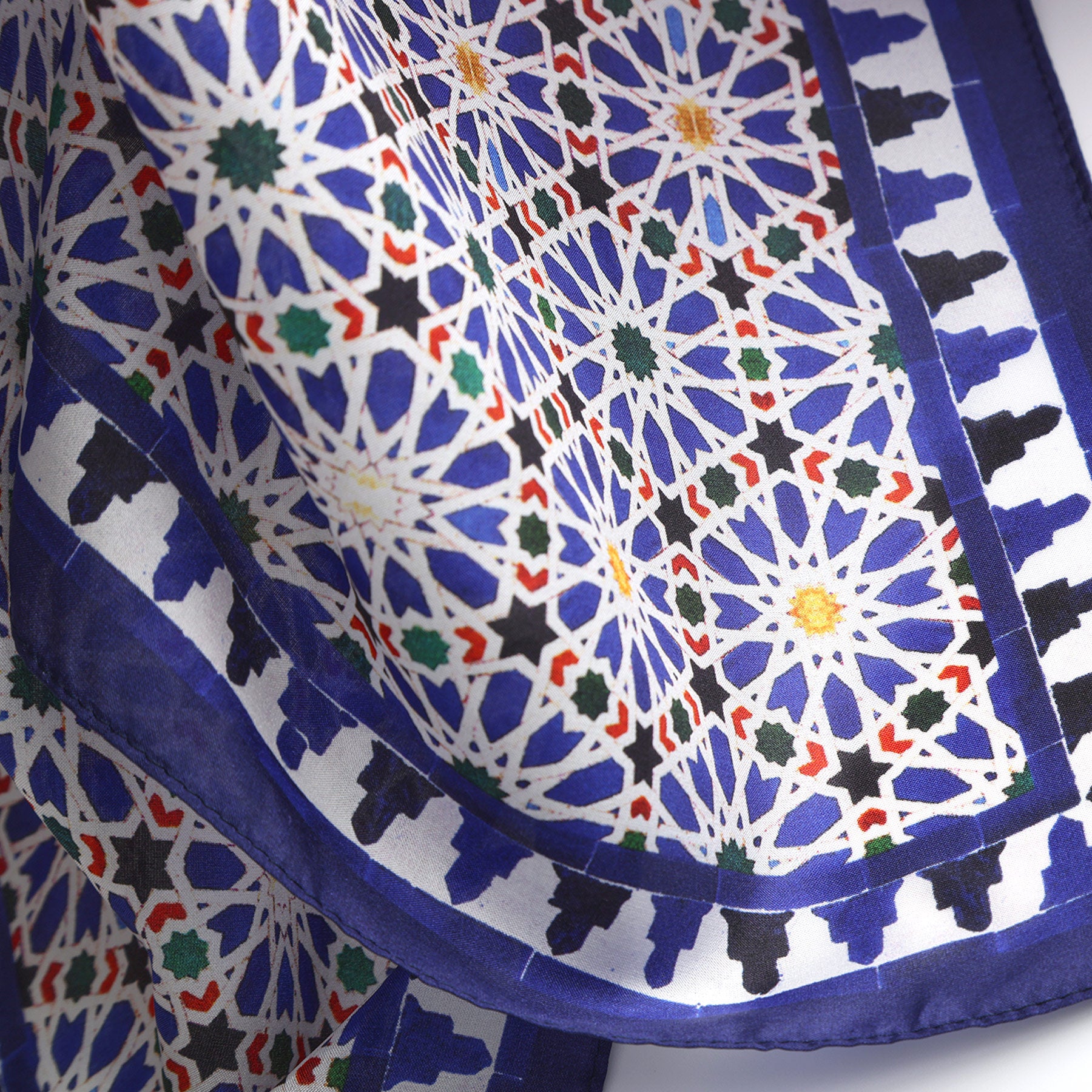 A close-up of the Blue Square Scarf Zellige v2 in silk, showcasing intricate geometric patterns in blue, white, yellow, green, and black with a bold blue border—drawing inspiration from traditional Islamic art and Spanish craftsmanship.