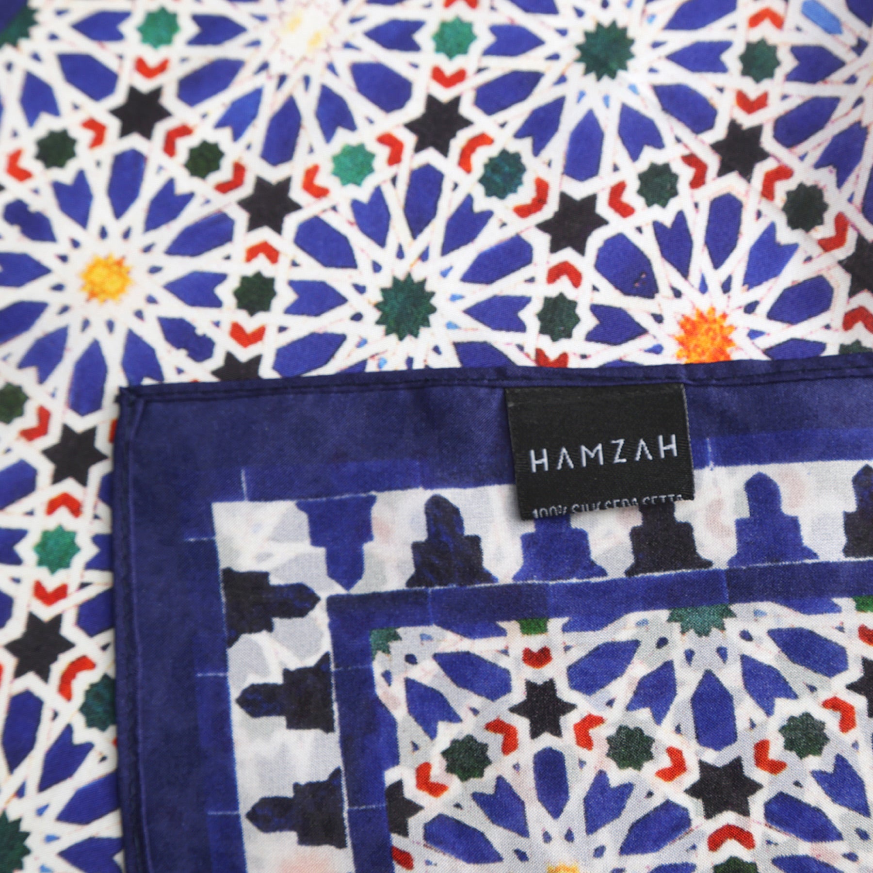 A close-up of the Blue Square Scarf Zellige v2, featuring a vibrant geometric Moroccan pattern in blue, white, green, and orange. The edge is blue-bordered with a sewn HAMZAH 100% COTTON label.