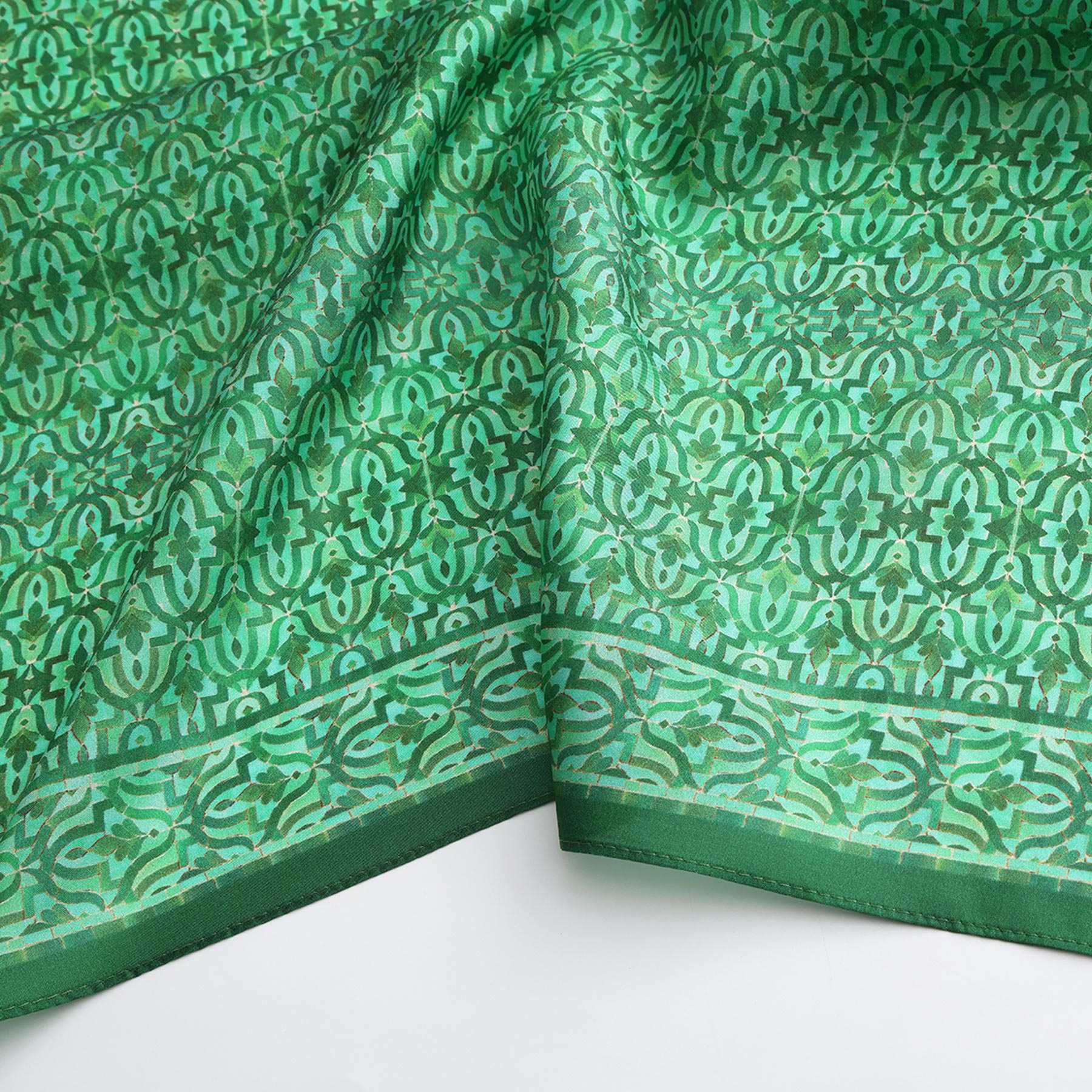 A close-up of the Green Square Silk Scarf Fihri, showcasing intricate Islamic art-inspired patterns and a decorative border on green silk, slightly folded and displayed against a white background.