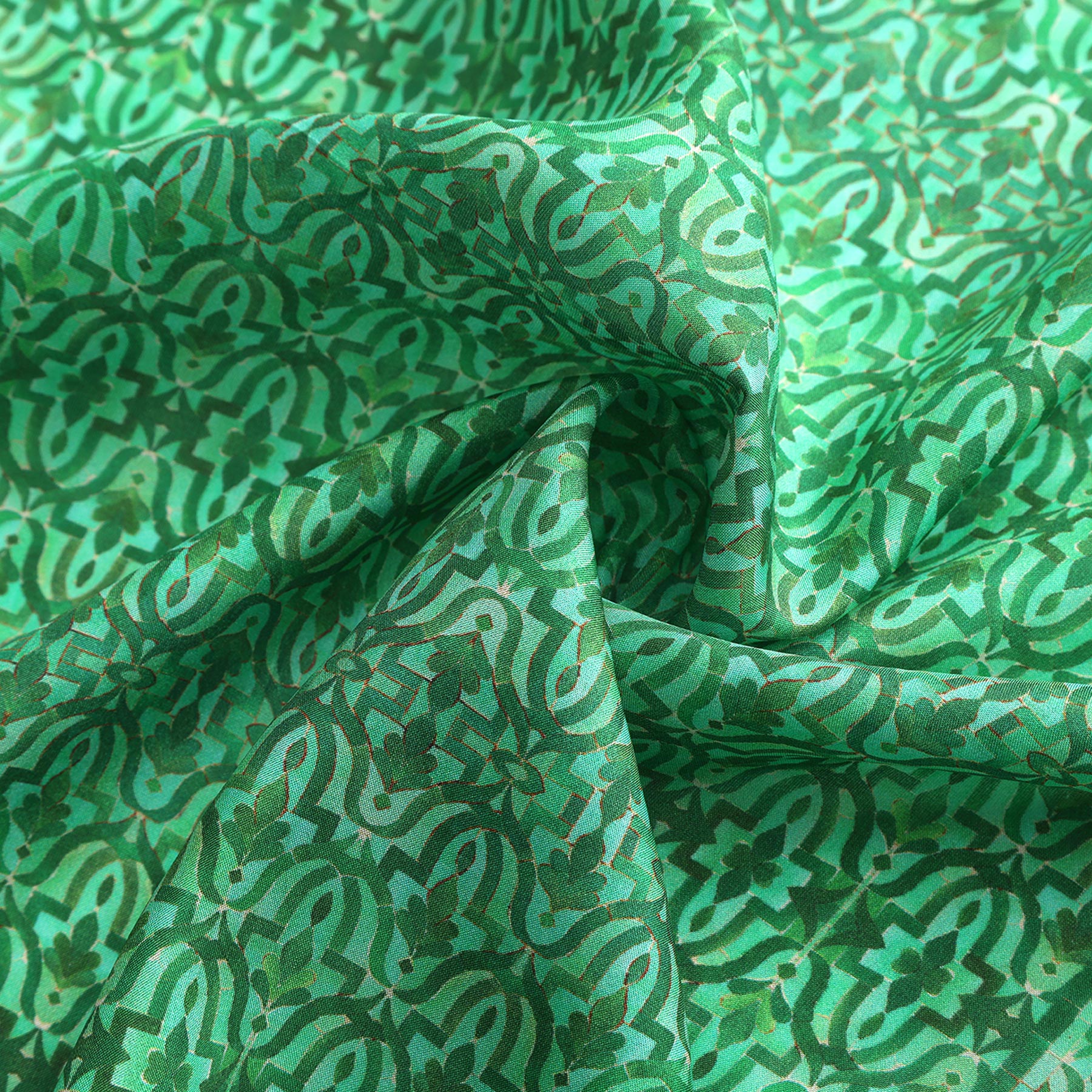 A close-up of the Green Square Silk Scarf Fihri shows its intricate geometric and floral pattern inspired by Islamic art, printed on green silk. The softly folded fabric reveals gentle creases and shadows, highlighting its textured look.