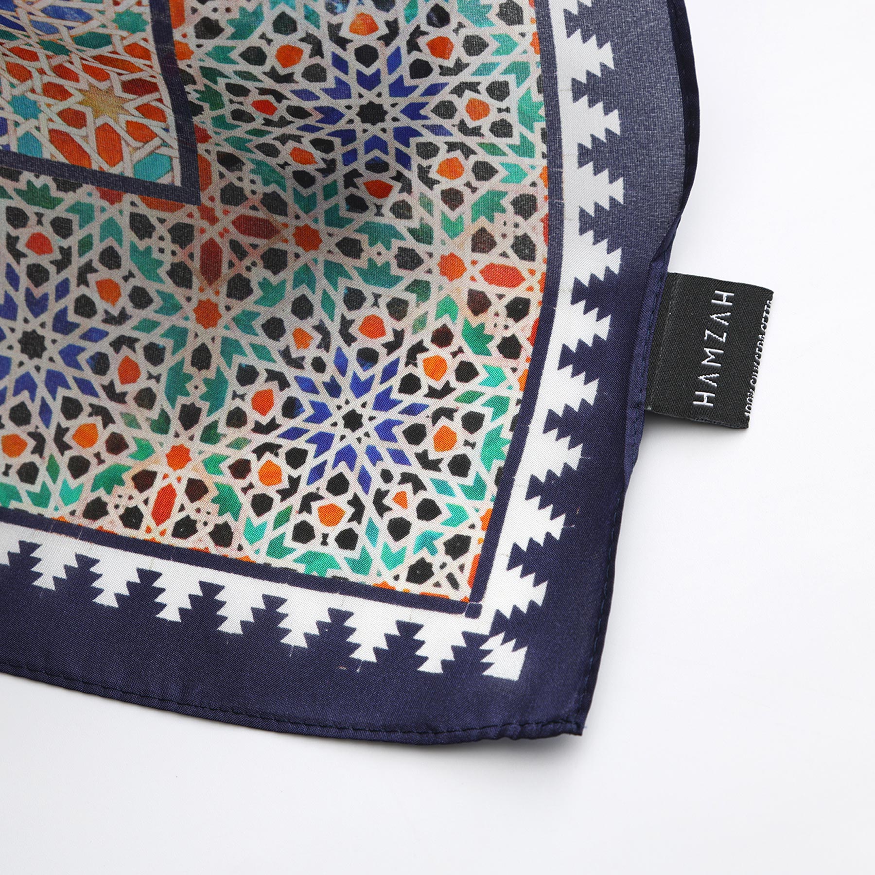 A close-up of the Square Scarf Blue Madiya, featuring intricate geometric patterns in blue, green, orange, and white with a navy border. A black tag with HAMZAH in white text is attached at the fabric’s edge.