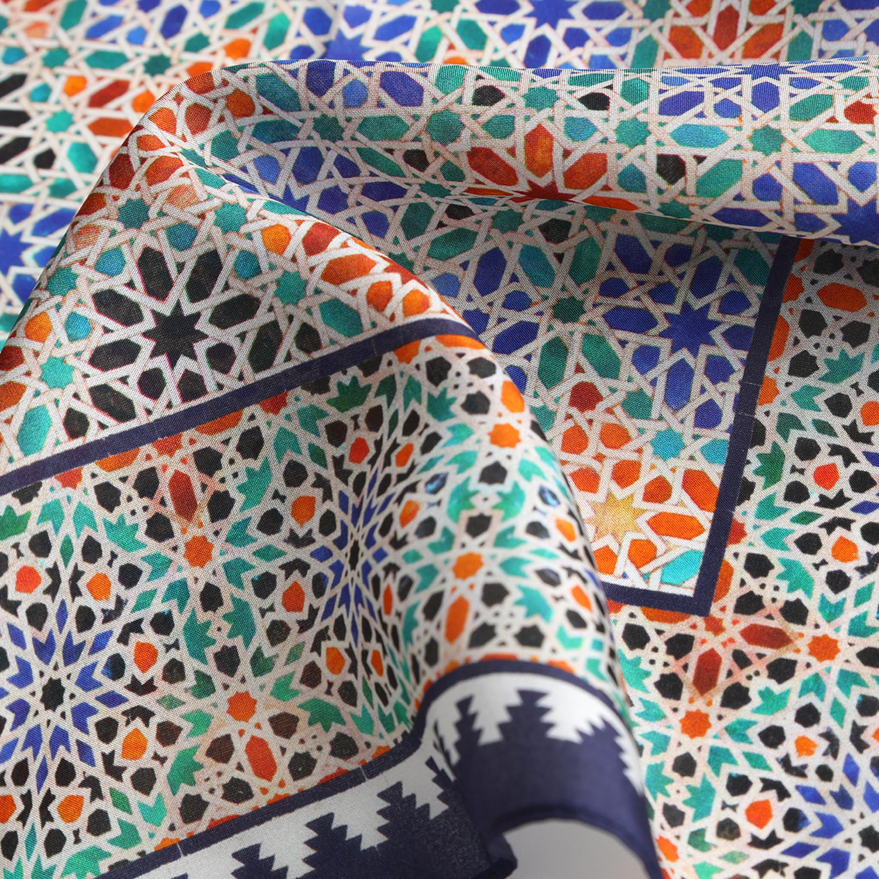 A close-up of the Square Scarf Blue Madiya, featuring intricate geometric patterns in orange, green, blue, and black inspired by Islamic art. The silk fabric is slightly folded, highlighting its texture and detailed design.