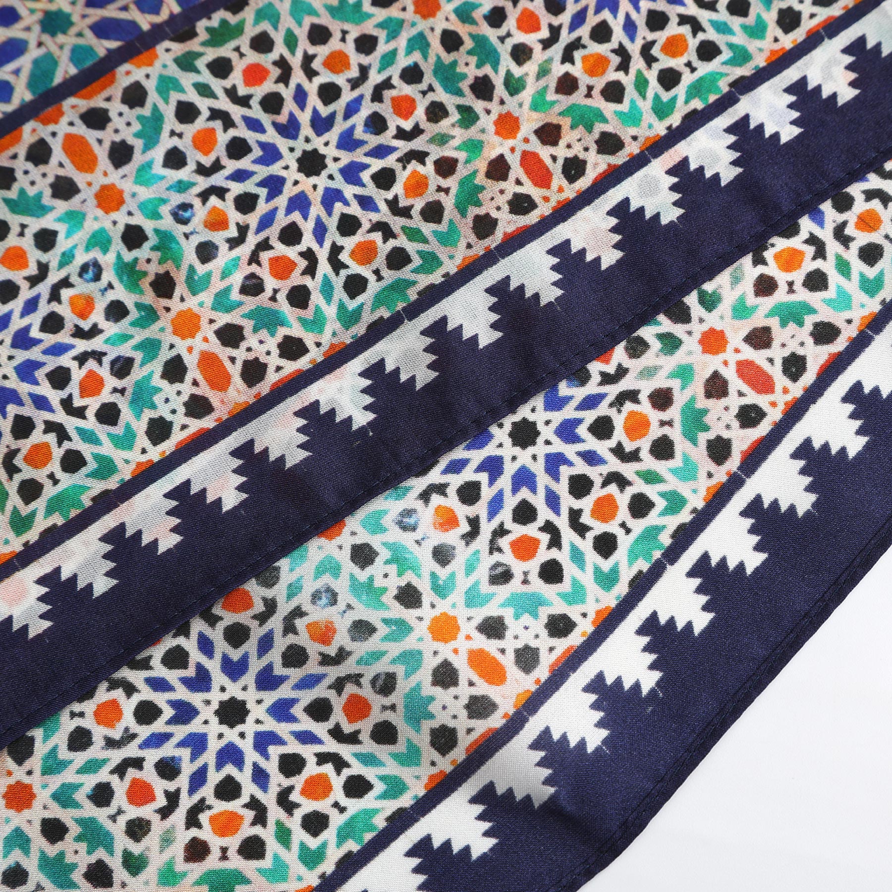 A close-up of the Square Scarf Blue Madiya shows intricate green, orange, and white geometric patterns on blue silk, bordered with a navy and white zigzag echoing Moroccan tile and Alhambra motifs.