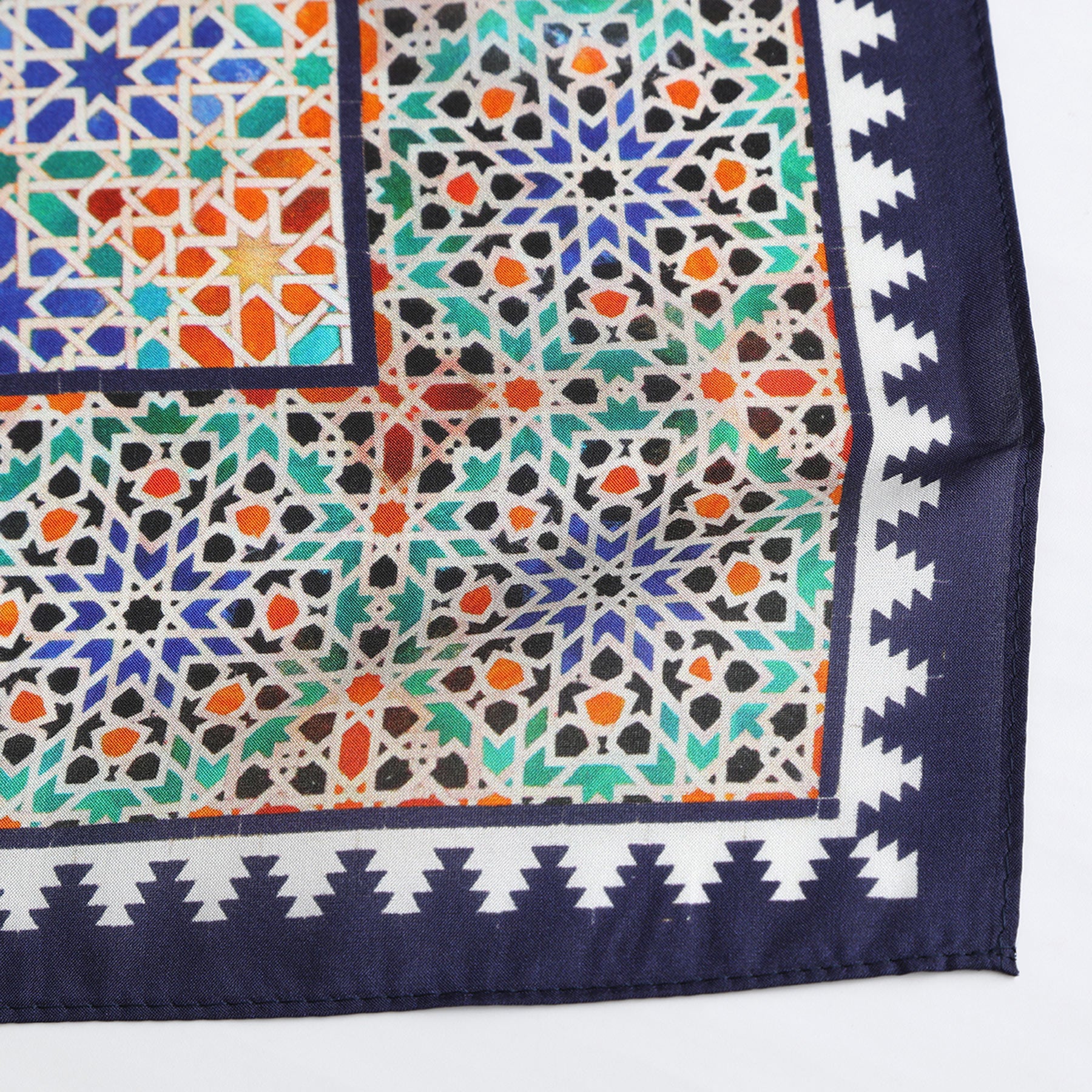 A close-up of the Square Scarf Blue Madiya, a silk piece featuring blue, green, orange, and white geometric patterns with a jagged navy and white border, inspired by majestic Alhambra Palace motifs.