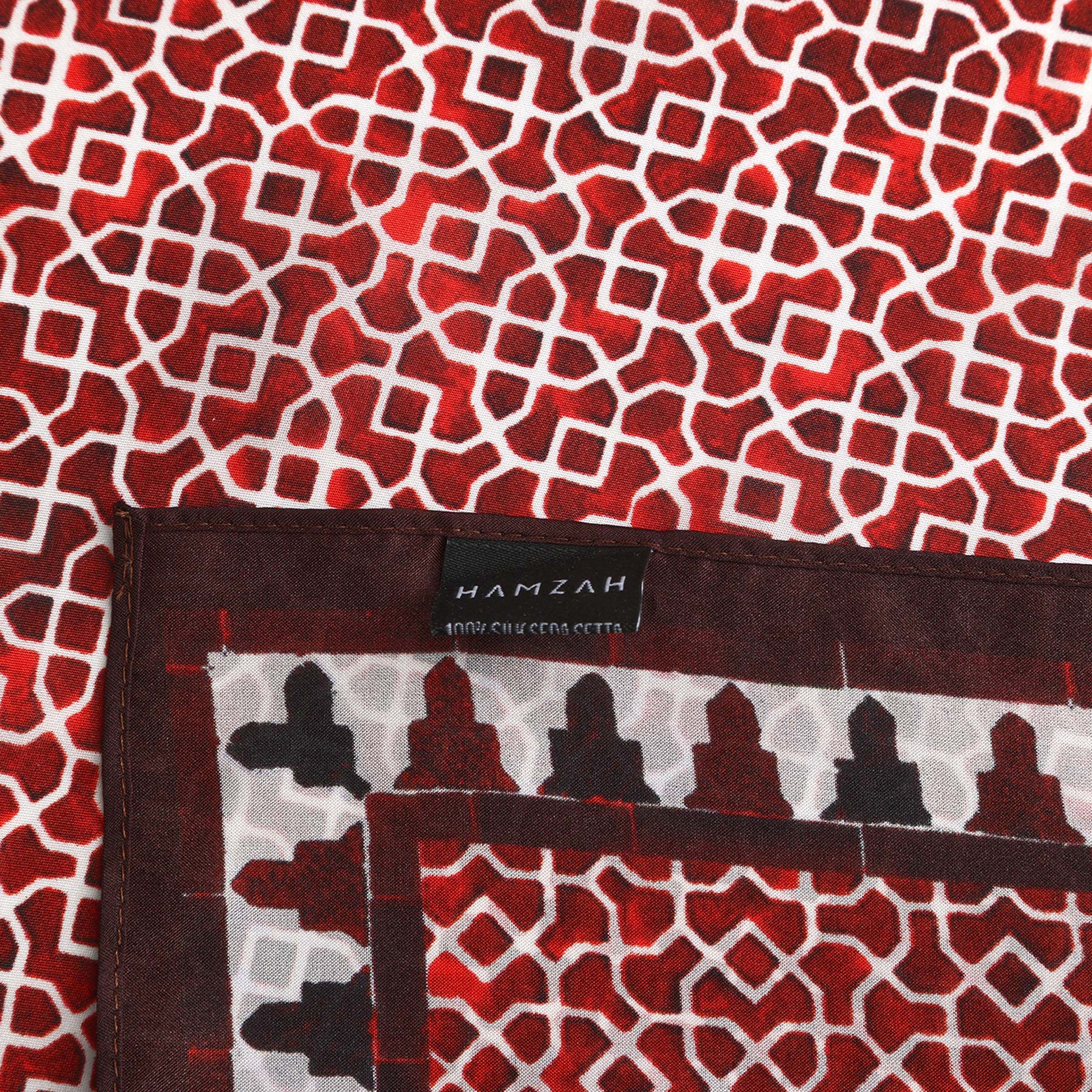 A close-up of the Red Square Scarf 100% Silk shows bold red, white, and black geometric designs inspired by Islamic art. The folded corner displays the black HAMZAH 100% SILK CERA SETTA label, highlighting its timeless elegance.