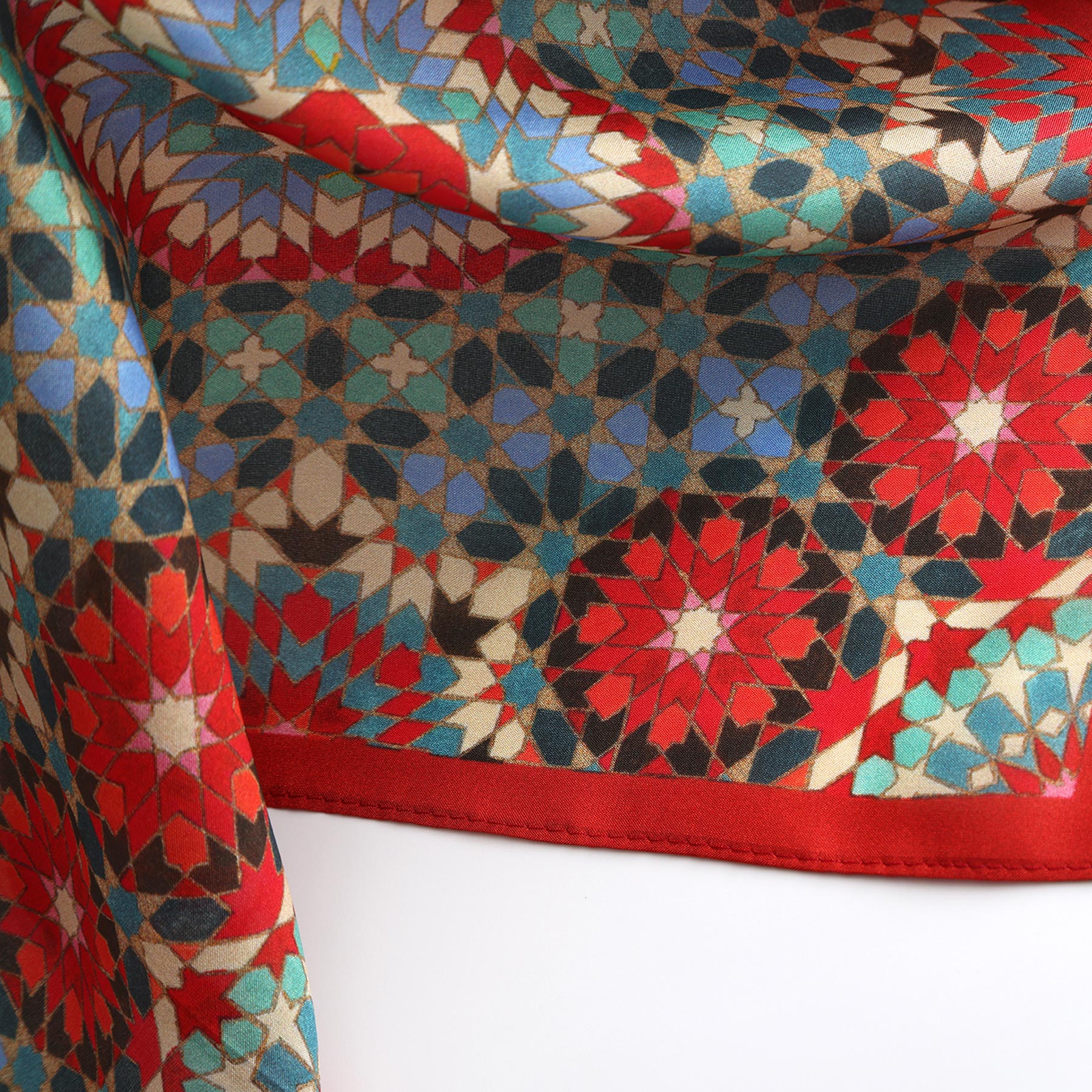 The Colorful Silk Neckerchief features a silky geometric mosaic inspired by Islamic art, with repeating star and floral shapes in red, blue, teal, black, and beige. The edge is finished with a neat red hem.