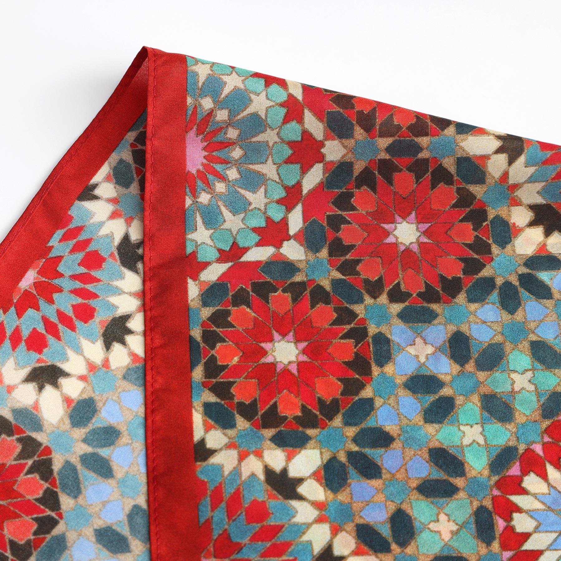 The Colorful Silk Neckerchief displays geometric, mosaic-inspired patterns in red, blue, black, and cream with a solid red border—a stylish nod to Islamic art—shown close-up on a white surface.