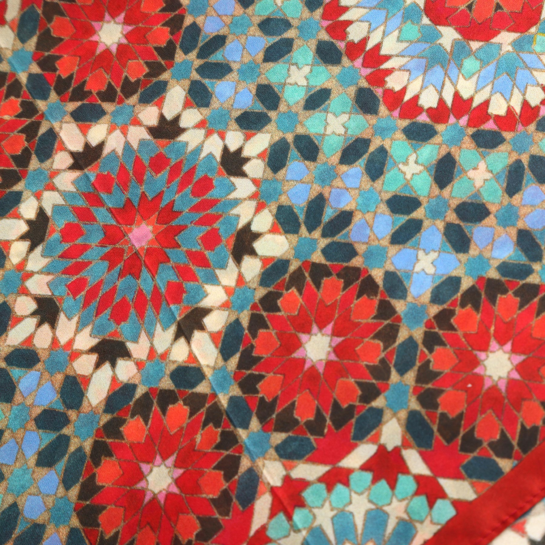 The Colorful Silk Neckerchief features a geometric mosaic inspired by Islamic art, with interlocking shapes in red, blue, white, brown, and teal forming symmetrical star patterns across the luxurious silk fabric.