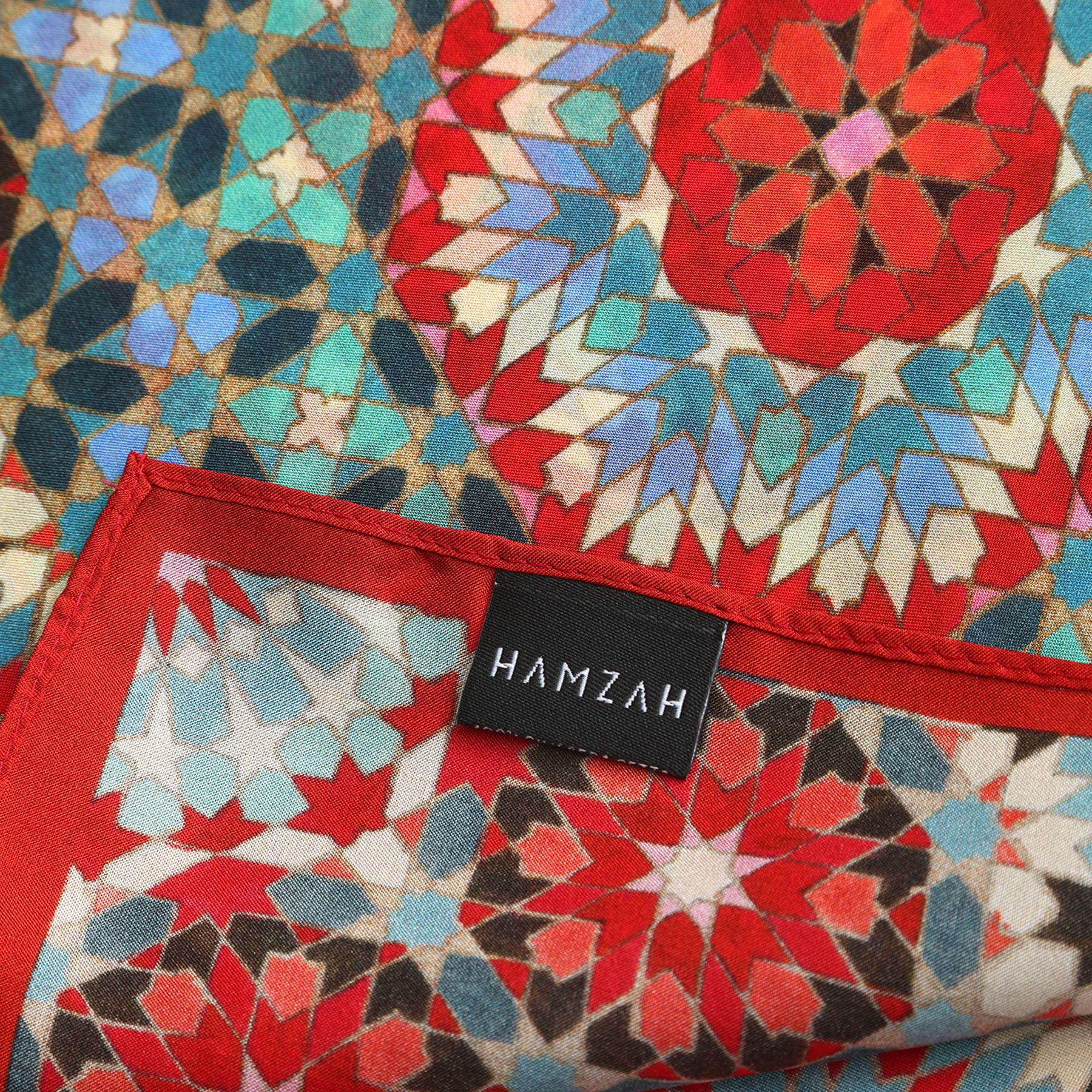 A close-up of the Colorful Silk Neckerchief, featuring geometric red, blue, and white patterns inspired by Islamic art, with a black HAMZAH label stitched in white.
