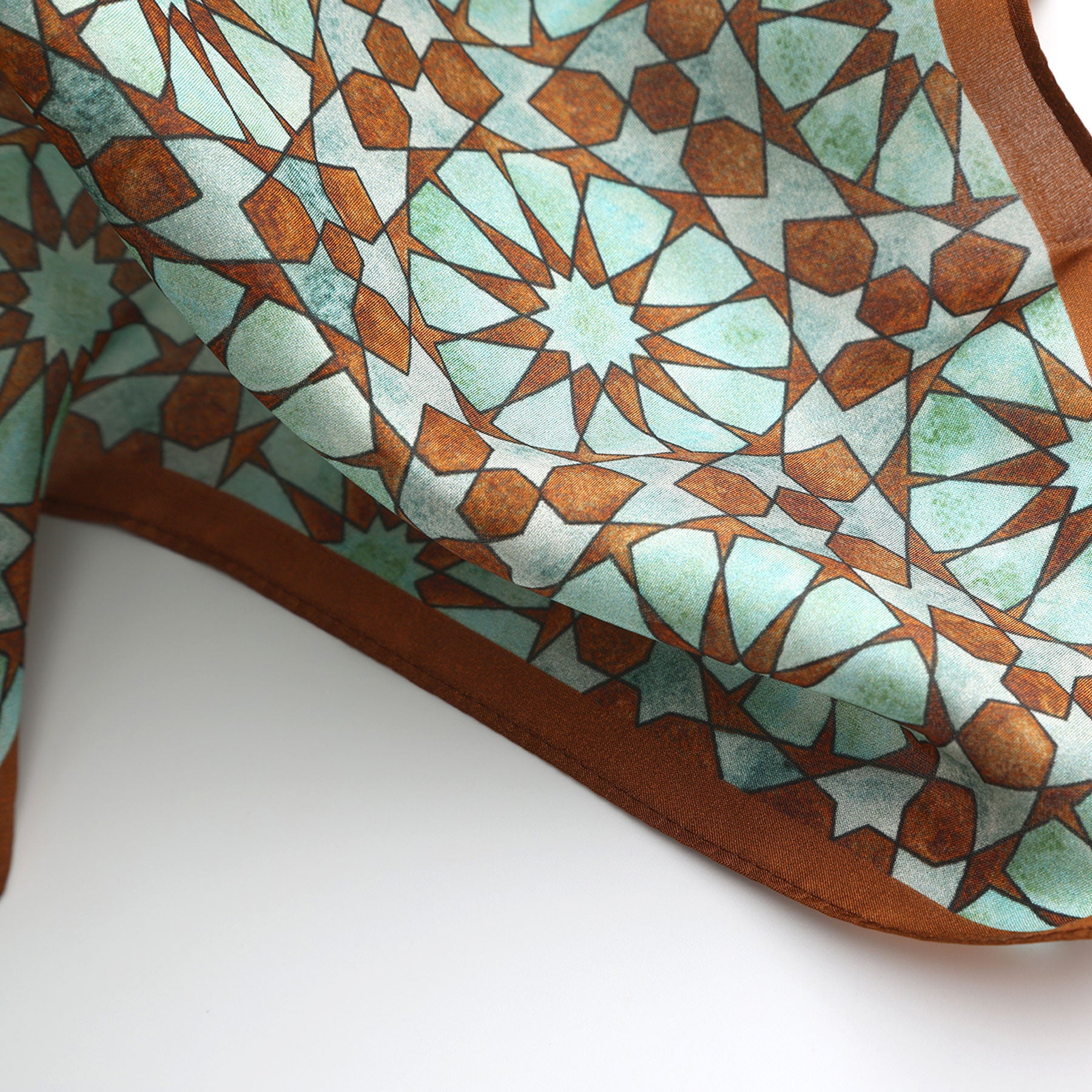 A close-up of the Brown Silk Neckerchief for Women showcases an intricate turquoise, brown, and white geometric pattern inspired by Islamic art, featuring repeating stars and polygons reminiscent of Spanish craftsmanship.