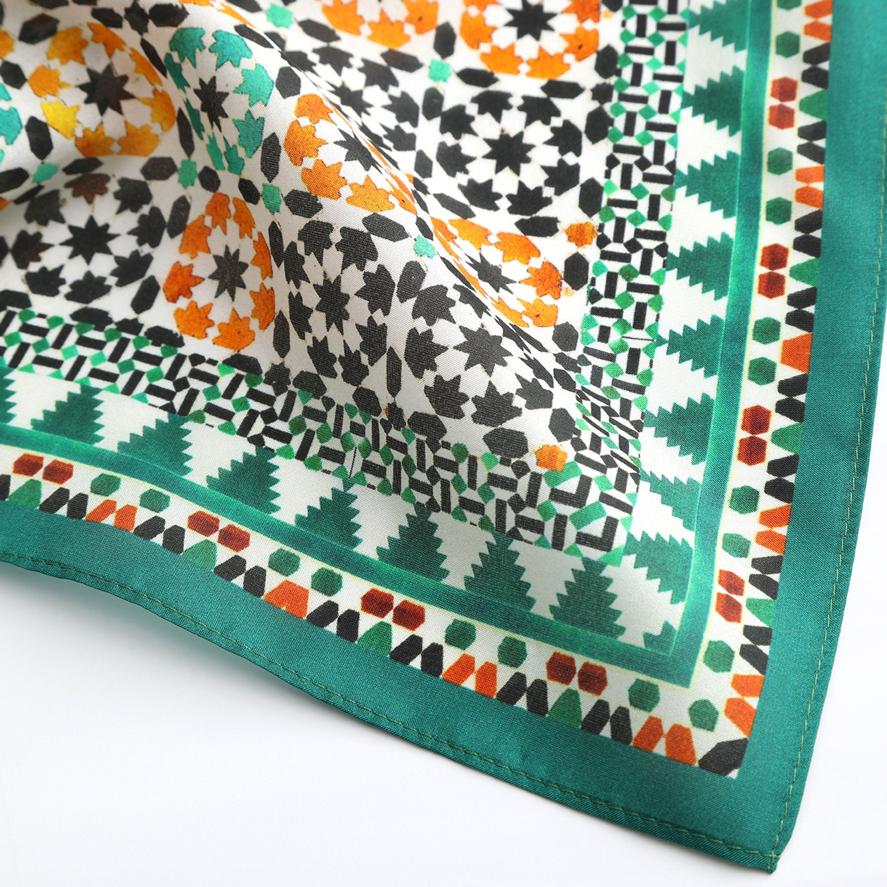 A close-up of the Green Square Silk Scarf Sabika with geometric patterns inspired by Islamic art in green, orange, black, and white. The scarf features repeating motifs, a green border, and a smooth, slightly shiny silk texture.