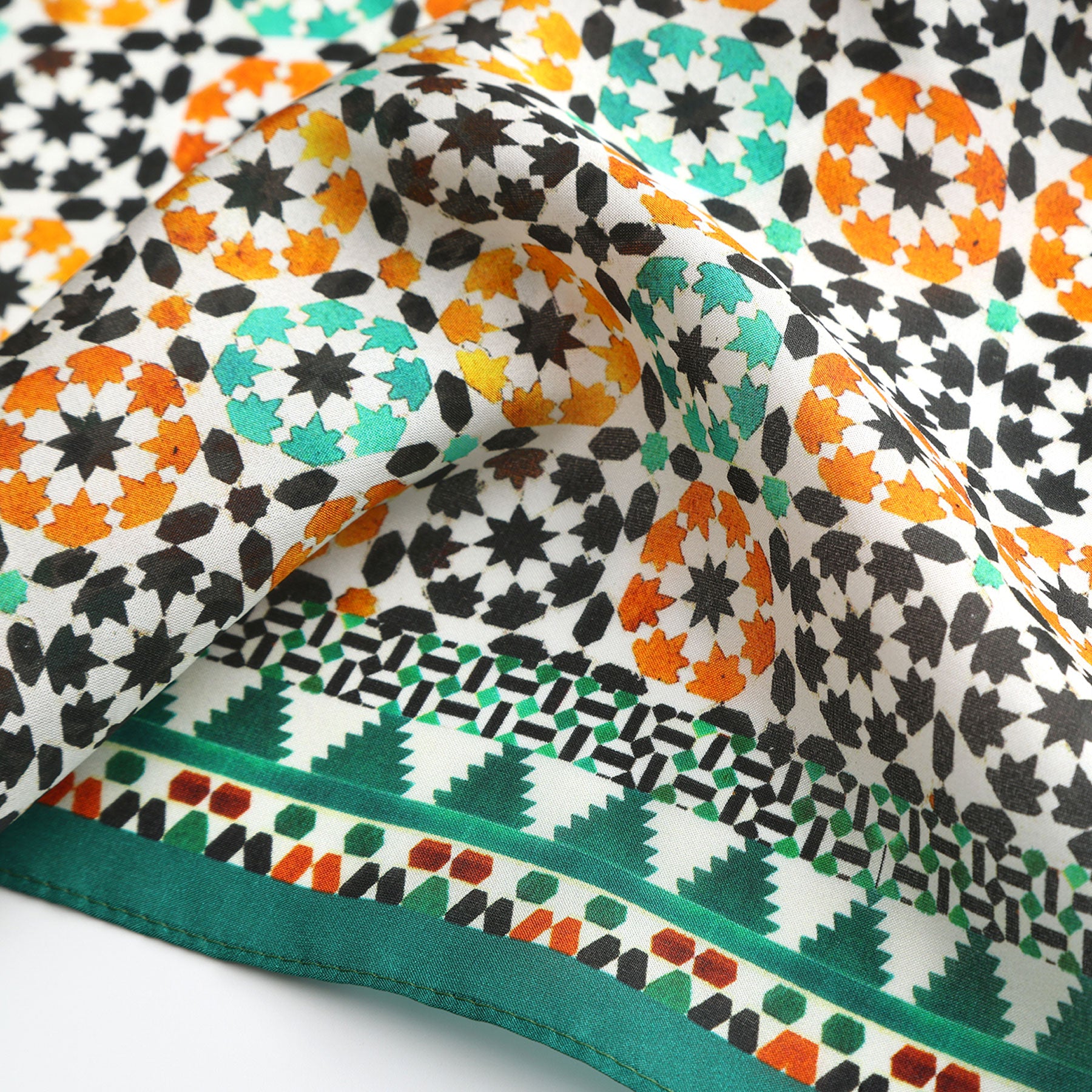 A close-up of the Green Square Silk Scarf Sabika shows soft, slightly gathered fabric with colorful geometric and floral Islamic art-inspired patterns in orange, green, teal, and black, accented by a distinct green and white triangular border.