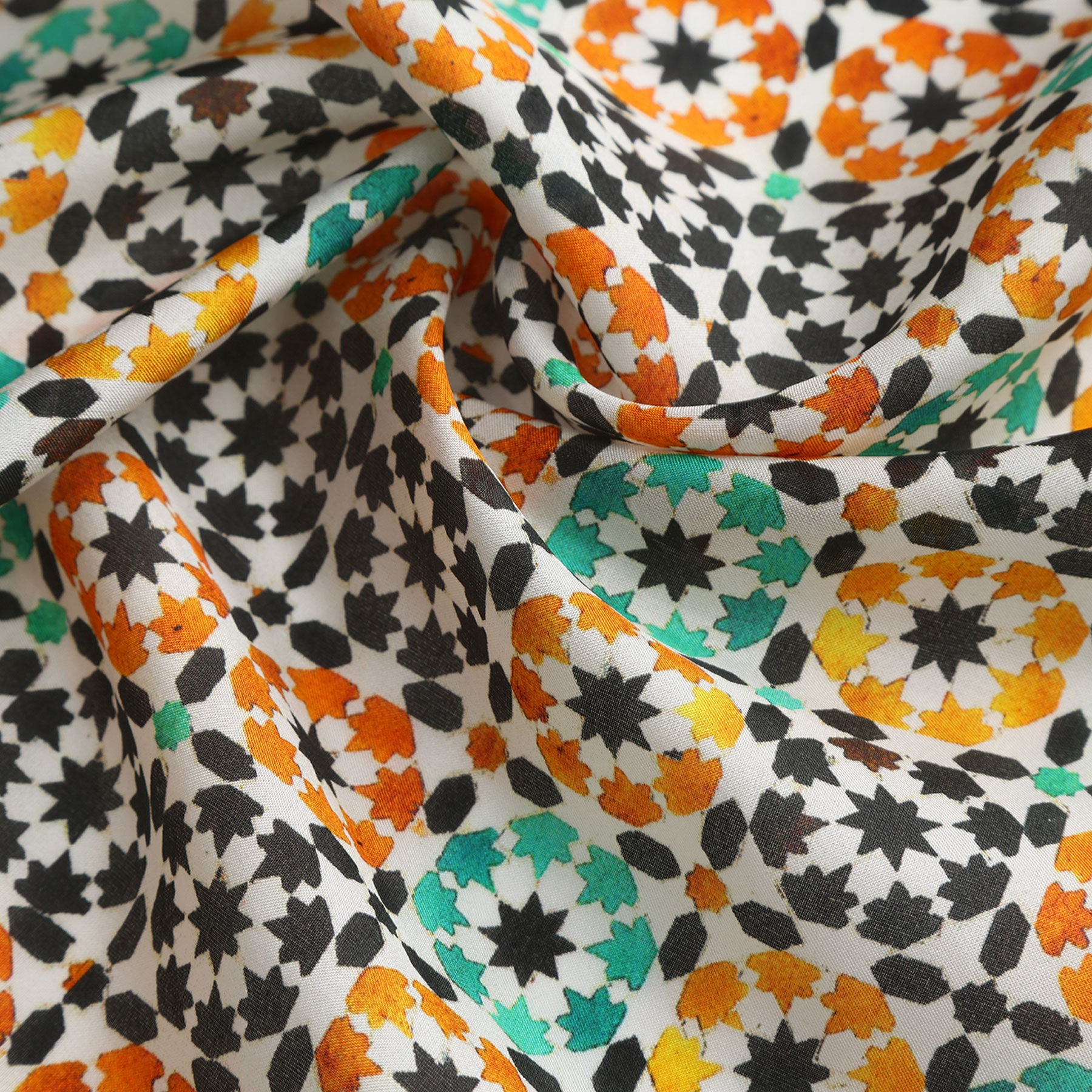 A close-up of the Green Square Silk Scarf Sabika, featuring intricate geometric designs in black, orange, yellow, and green on white. Inspired by Islamic art, the softly folded silk creates gentle shadows and rich texture.