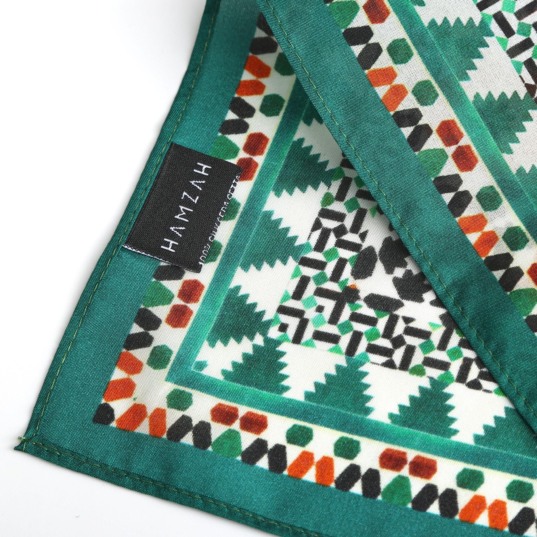 A close-up of the Green Square Silk Scarf Sabika shows green-bordered fabric with geometric patterns in green, black, and orange inspired by Islamic art. A black label reads HAMZAH Hand crafted. The folded scarf highlights its intricate design.