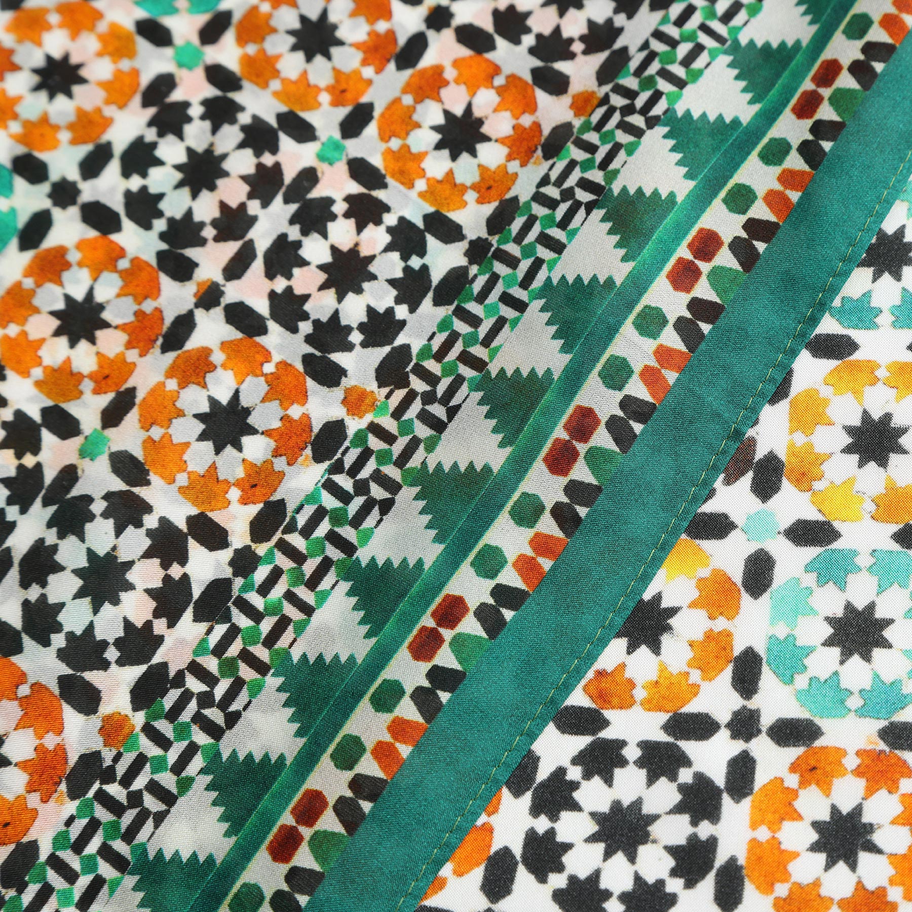 Close-up of the Green Square Silk Scarf Sabika, showcasing geometric patterns in orange, black, white, and green with a diagonal green border. The design evokes Islamic art motifs and highlights exquisite Spanish craftsmanship.