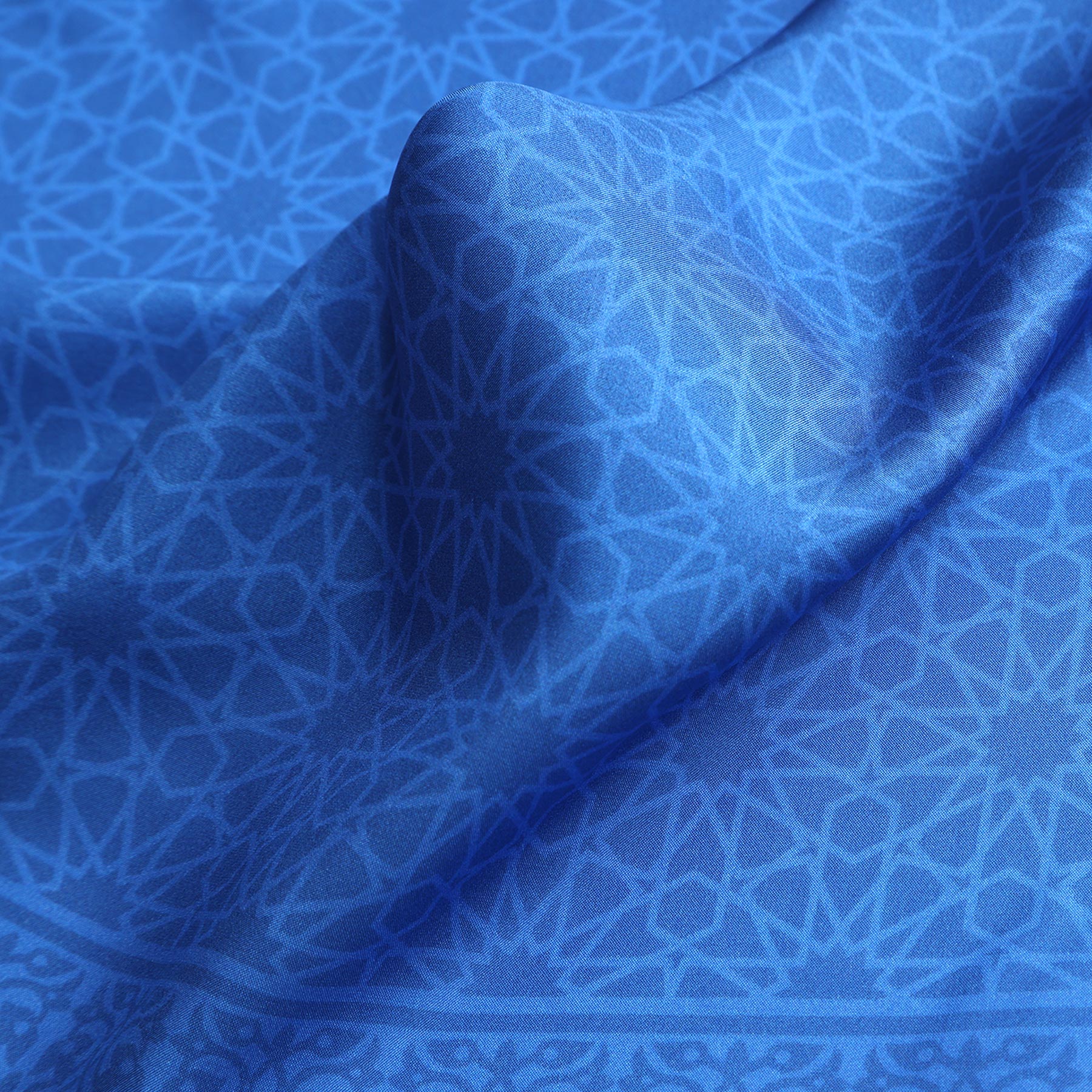 The Blue Square Silk Scarf Dalila v1 features a close-up of intricate blue geometric patterns, inspired by Islamic art motifs from the Alhambra Palace. Repeating star shapes and interlocking lines create a subtle texture on its soft, lustrous silk fabric.