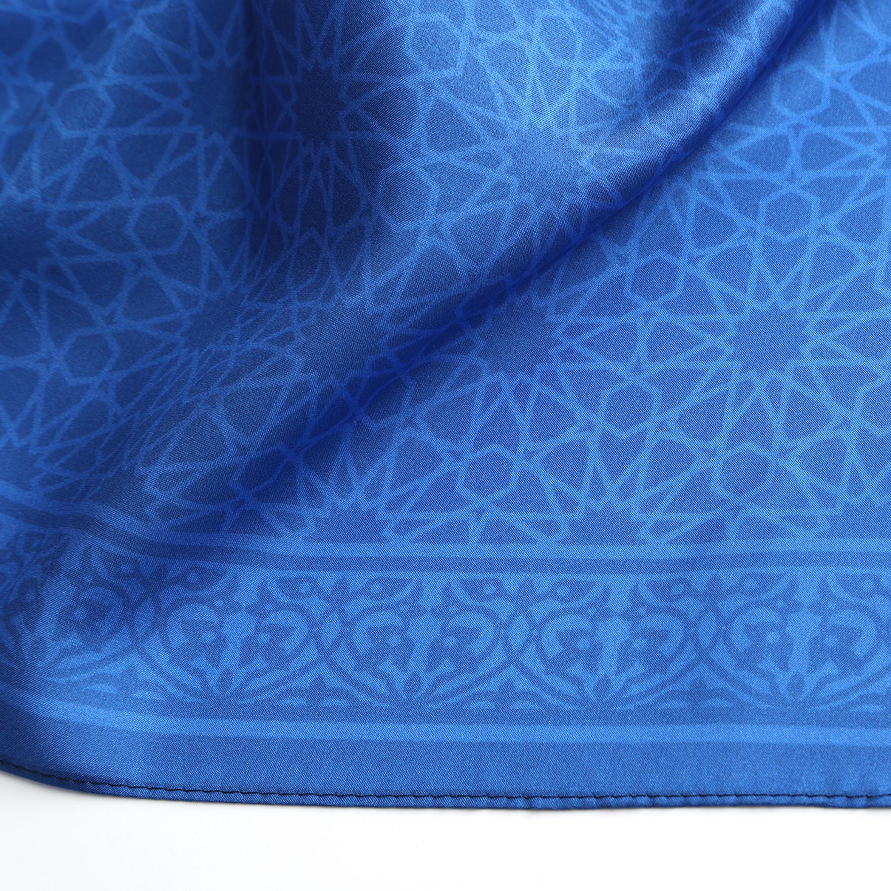 A close-up of the Blue Square Silk Scarf Dalila v1 shows intricate geometric and floral patterns inspired by Islamic art, with a decorative border. The design features a subtle sheen and symmetrical, repeating motifs.