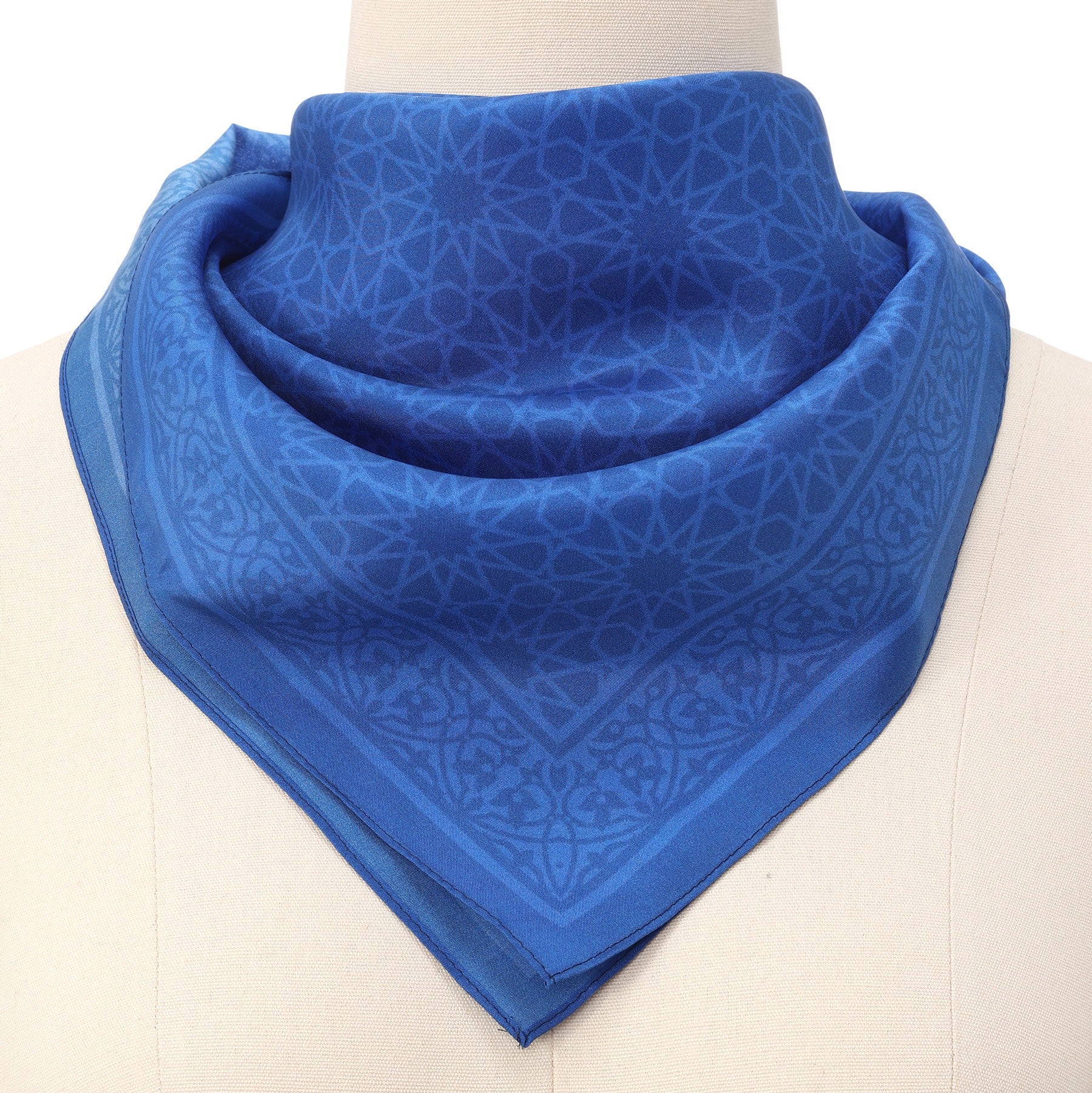 A mannequin showcases the Blue Square Silk Scarf Dalila v1, featuring a subtle geometric pattern inspired by Islamic art, folded into a triangle and elegantly draped around the neck.