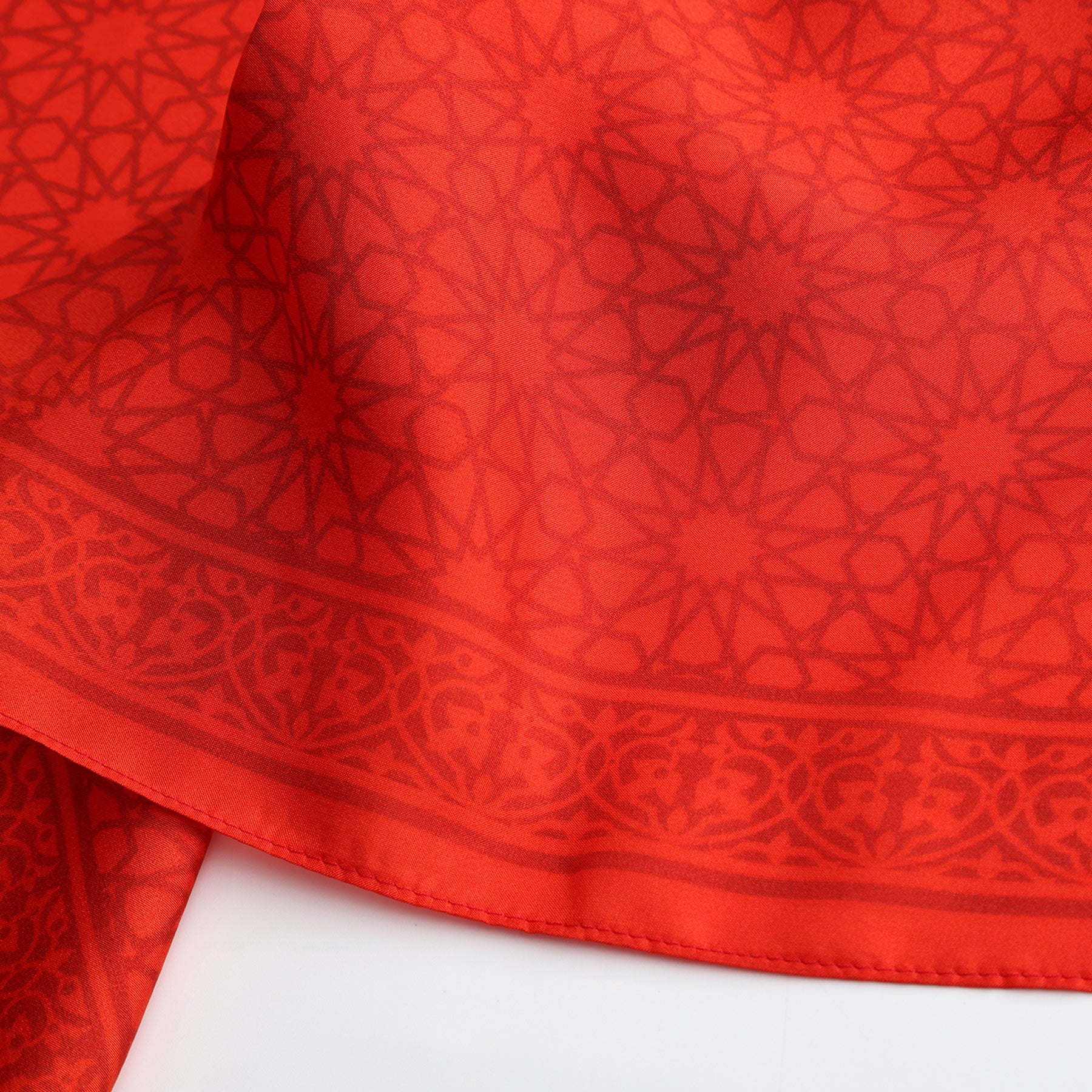 A close-up of the Red Square Scarf Dalila v2, crafted from 100% silk, showcases its intricate Islamic-inspired geometric and floral patterns with a decorative border, artfully draped on a white surface.