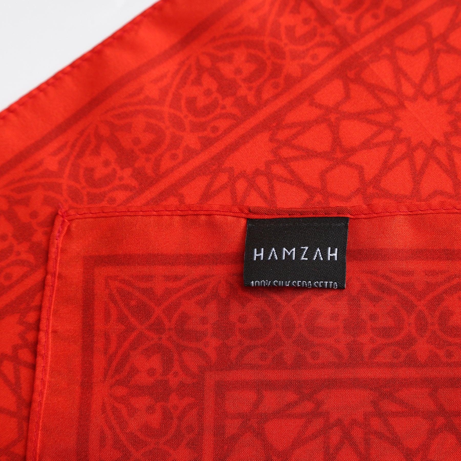 Close-up of the Red Square Scarf Dalila v2 - 100% Silk, featuring a red geometric and floral pattern inspired by Islamic art, with a black rectangular HAMZAH label stitched on.