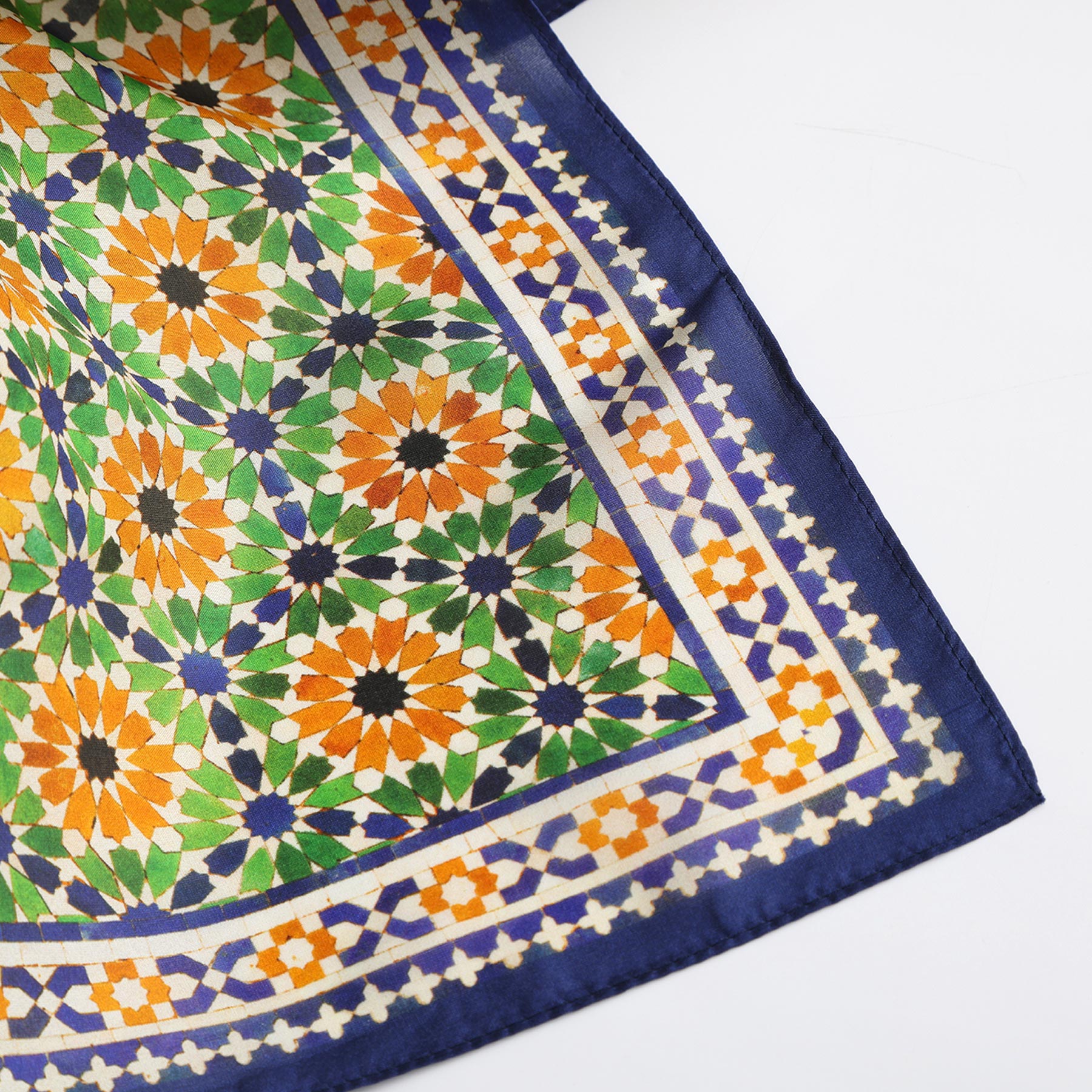 A close-up of the Islamic Art Print Square Silk Scarf Nur shows a geometric mosaic pattern in green, orange, black, and white with a blue and cream border on white, inspired by Islamic art.