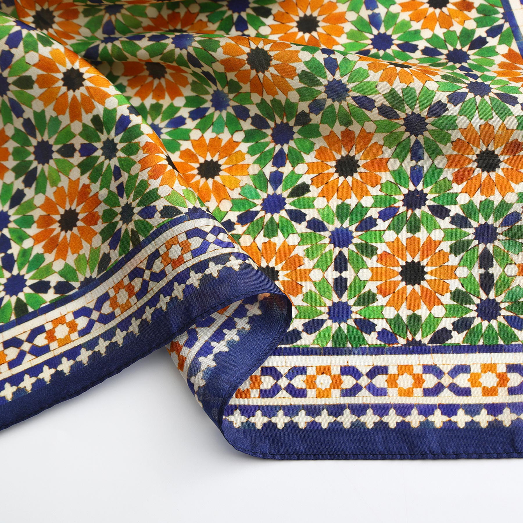 The Islamic Art Print Square Silk Scarf Nur features a geometric mosaic design in green, orange, white, and dark blue with a decorative border and folded corner, inspired by Islamic art and displayed on a white surface.