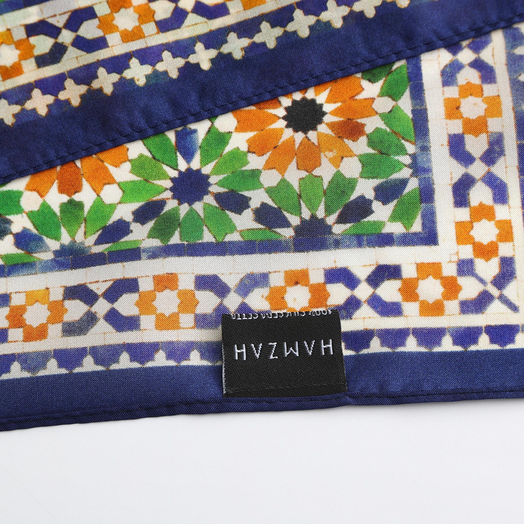 A close-up of the Islamic Art Print Square Silk Scarf Nur, showcasing green, orange, and blue geometric designs. A black label marked “HAMZAH” is stitched to the edge, reflecting refined Spanish craftsmanship.