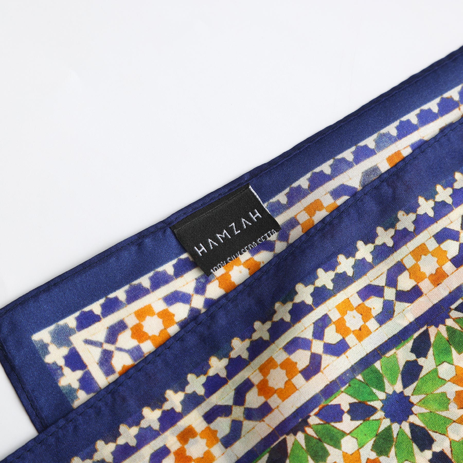 A close-up of the Islamic Art Print Square Silk Scarf Nur, showcasing geometric designs in blue, orange, green, and white with a dark blue border and a black “HAMZAH” label sewn on the edge.