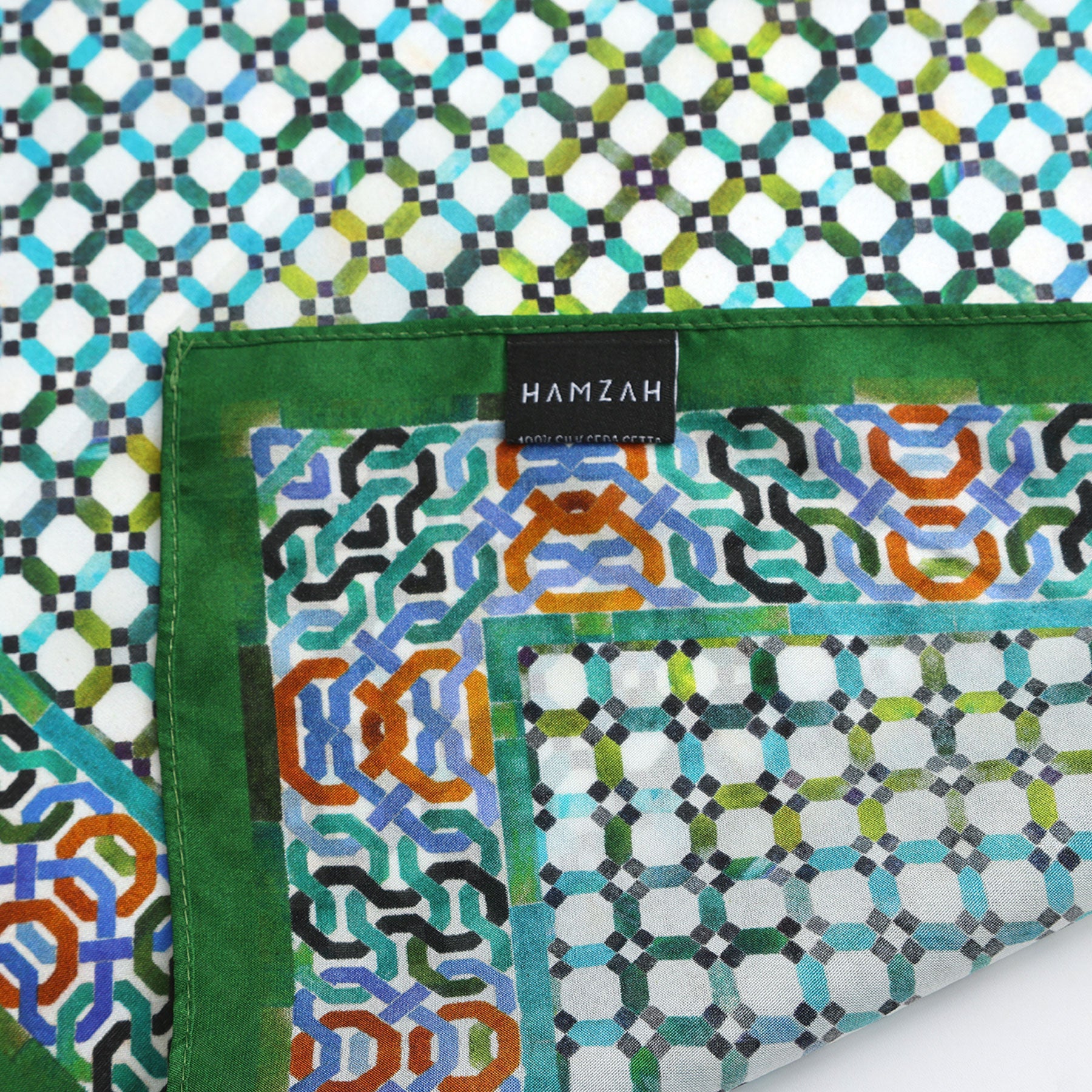 A close-up of the Coloful Neckerchief 100% Silk, featuring intricate geometric patterns in green, blue, black, and orange. The neckerchief is partially folded to reveal a HAMZAH label and echoes traditional Islamic art.