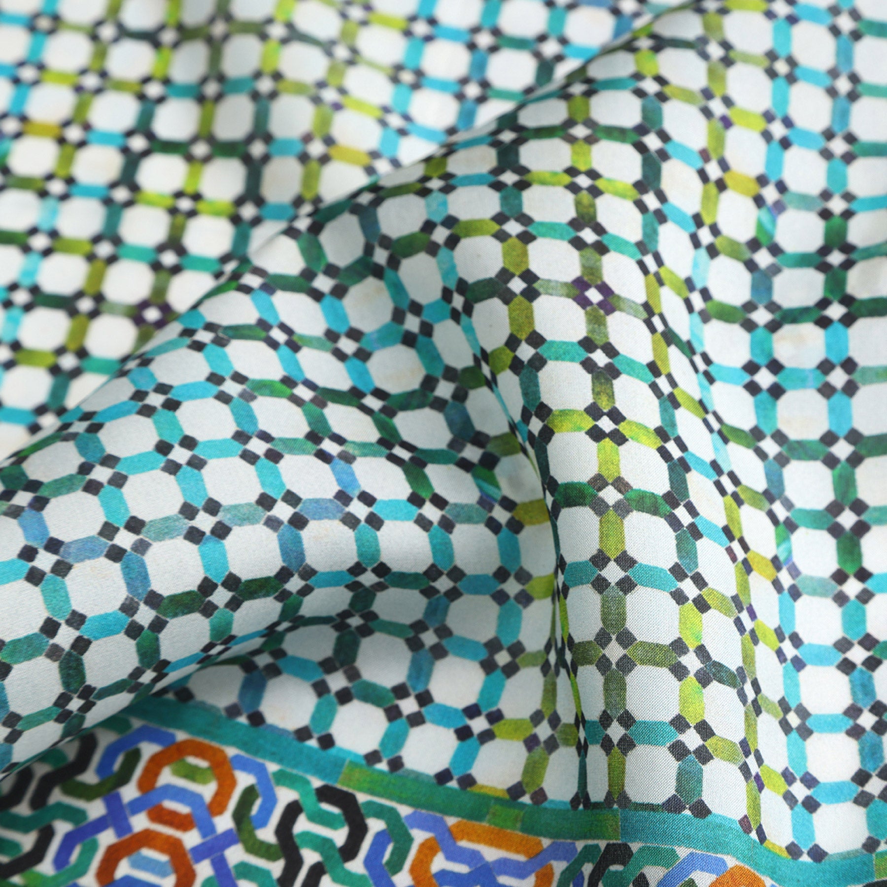 A close-up of the Coloful Neckerchief 100% Silk, showcasing a geometric Alhambra Palace print in blue, green, black, and white with interlocking hexagons, circles, and a colorful border along the bottom edge.