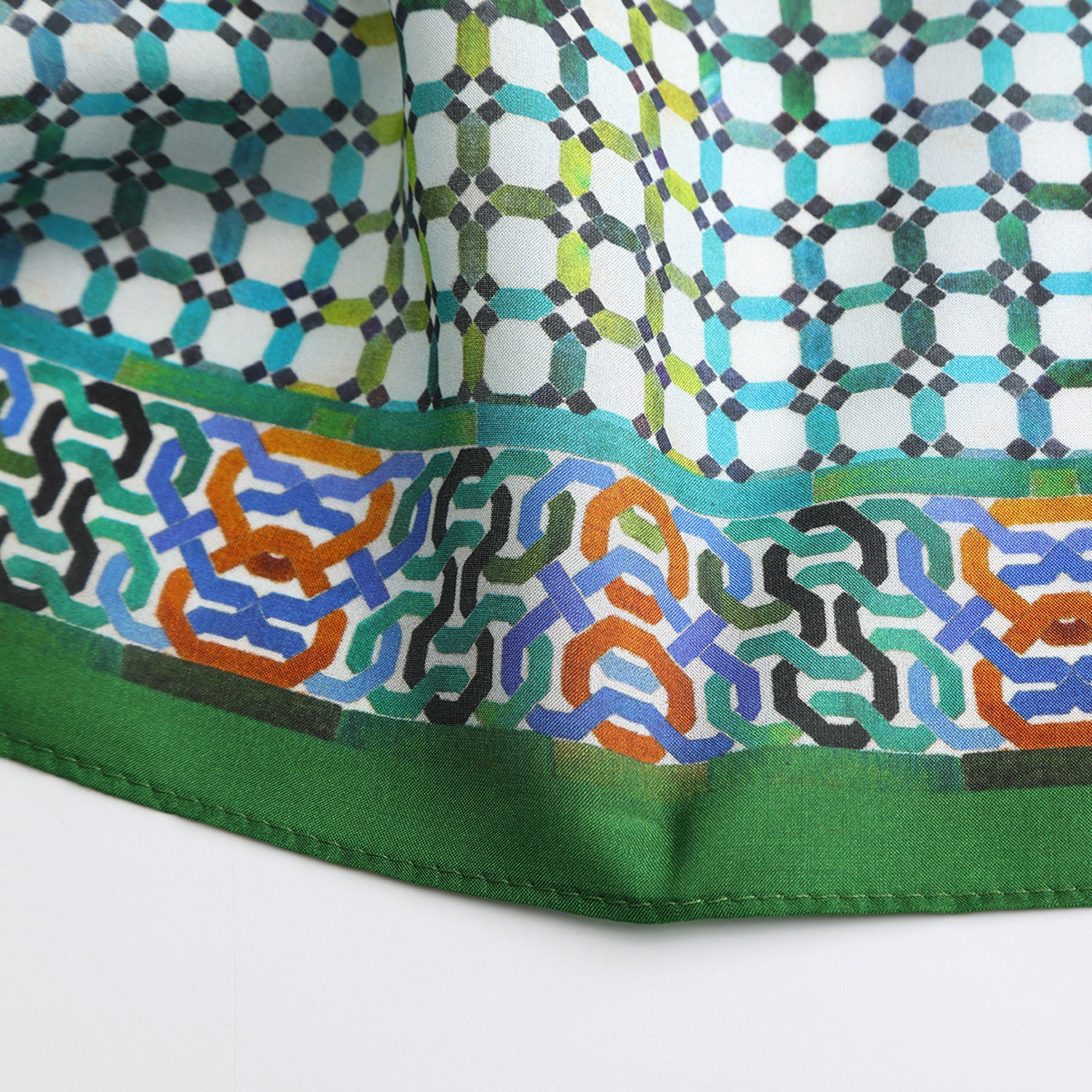 The Coloful Neckerchief 100% Silk features a vibrant geometric pattern in blue, green, black, and orange with a solid green border. Inspired by Islamic art, its design shows interlocking shapes and repeating motifs.
