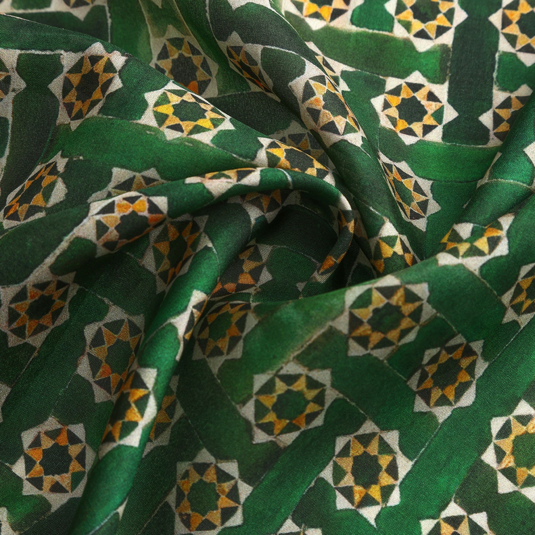A close-up of the Green Square Scarf Silk shows its Alhambra-inspired geometric pattern with yellow, black, and white star shapes and interlocking green lines. The slightly wrinkled silk fabric highlights the intricate, eye-catching design.